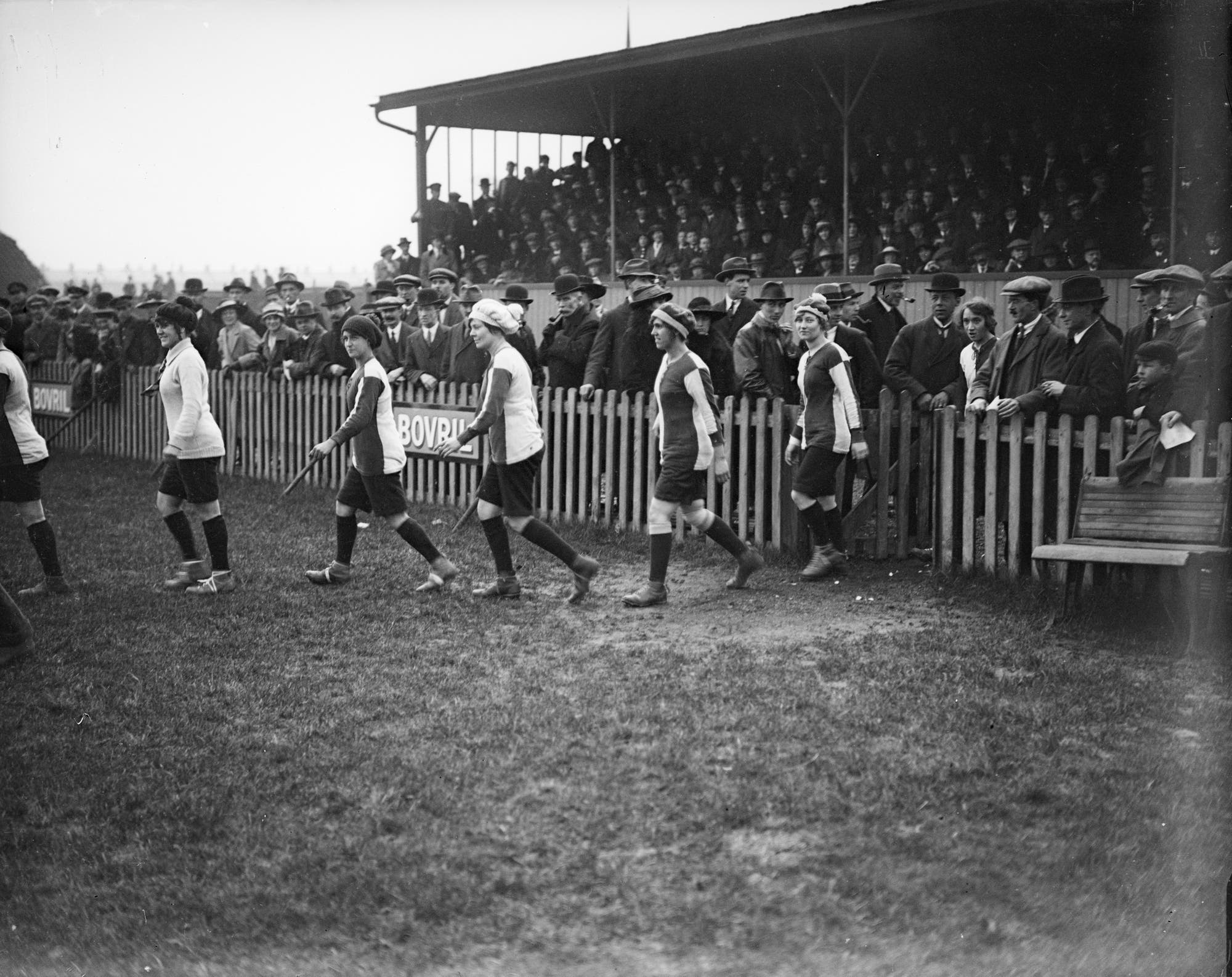 Eine Gruppe von Frauen in Sportkleidung geht auf einem Fußballfeld entlang, während eine große Menschenmenge auf der Tribüne zuschaut. Die Frauen tragen Trikots und Stulpen, einige mit Kopfbedeckungen. Im Hintergrund ist ein Zaun mit einem Werbeschild für "Bovril" sichtbar. Die Zuschauer tragen überwiegend Hüte und Mäntel, was auf eine historische Aufnahme hindeutet. Die Szene vermittelt eine sportliche Veranstaltung mit regem Zuschauerinteresse.