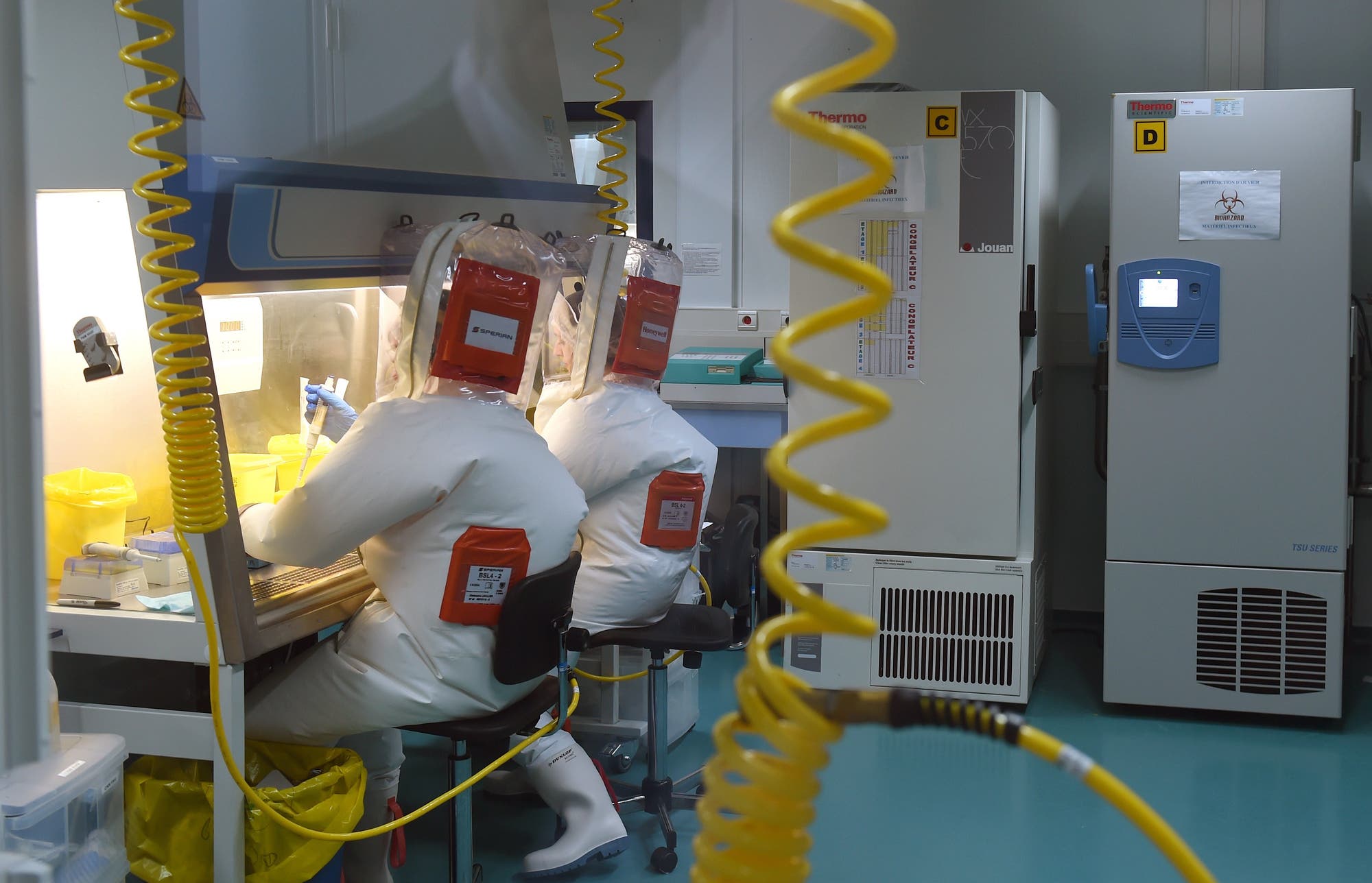 Zwei Personen in Schutzanzügen arbeiten in einem Labor an einer Sicherheitswerkbank. Sie tragen Vollschutzanzüge mit Atemgeräten und hantieren mit Laborutensilien. Im Hintergrund sind Kühlschränke mit Sicherheitskennzeichnungen zu sehen.