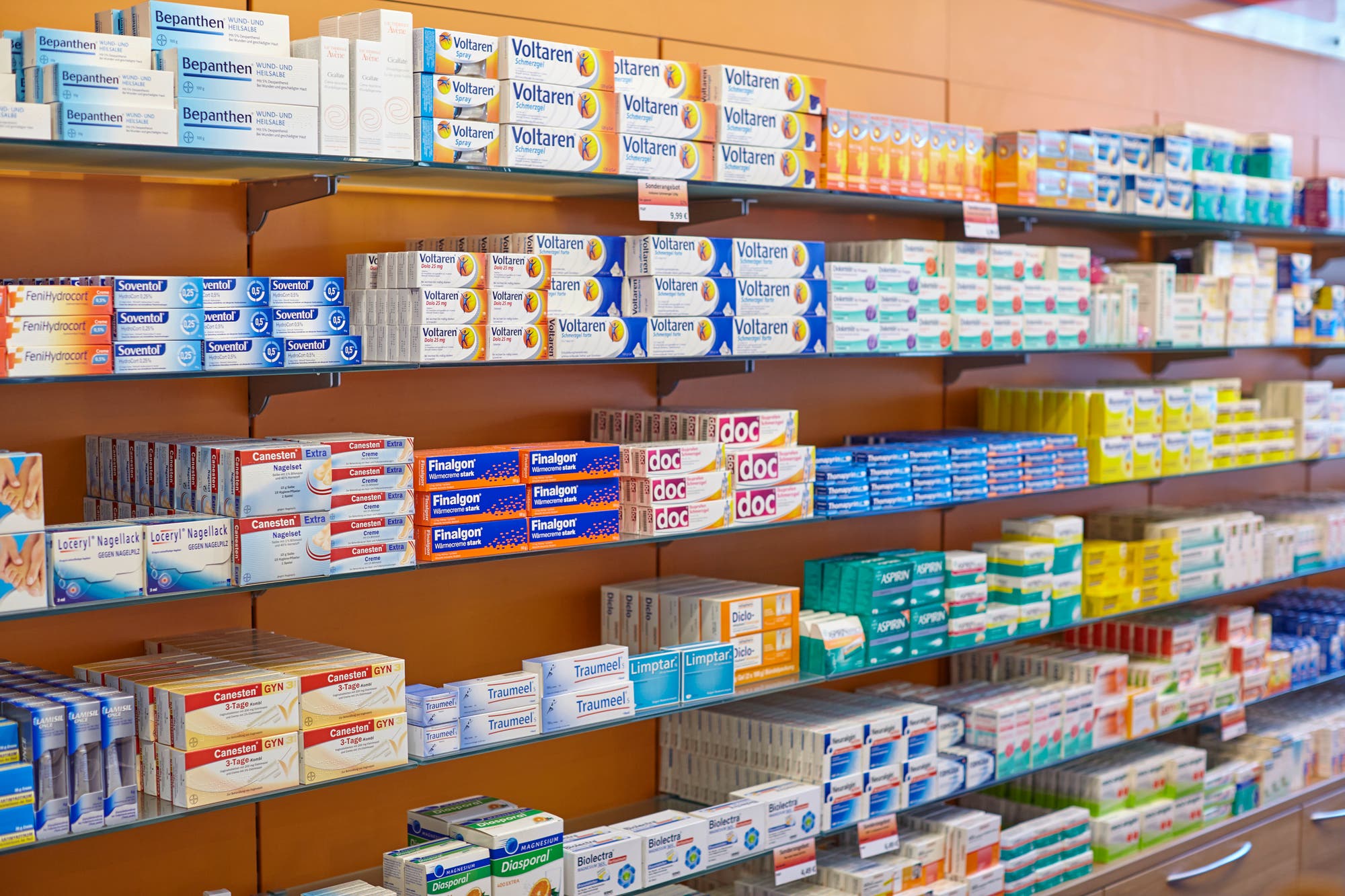 Regale in einer Apotheke, gefüllt mit verschiedenen Medikamenten und Salben. Zu sehen sind Marken wie Voltaren, Bepanthen und Fenistil. Die Produkte sind ordentlich in Reihen angeordnet. Im Vordergrund sind Schilder mit Preisen sichtbar. Die Umgebung wirkt sauber und gut organisiert, typisch für eine Apothekenauslage.