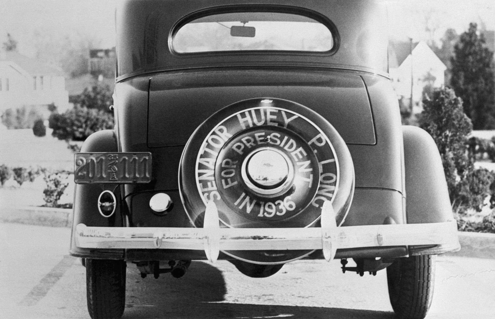 Rückansicht eines Autos aus den 1930er Jahren, geparkt auf einer Straße. Auf dem Reserverad ist der Text "Senator Huey P. Long for President in 1936" zu sehen. Das Nummernschild zeigt die Zahlen "211111". Im Hintergrund sind unscharf Bäume und Häuser zu erkennen.