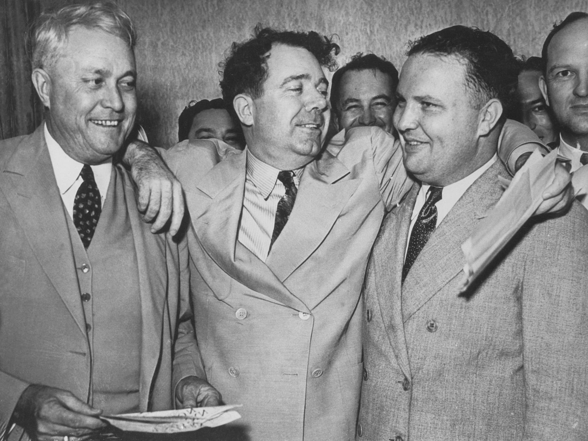 Eine Gruppe von Männern in Anzügen steht in bester Laune eng beieinander. Der Mann in der Mitte – Long – hat die Arme auf und um die Schultern der beiden anderen gelegt. Die Politiker feiern augenscheinlich einen Sieg.