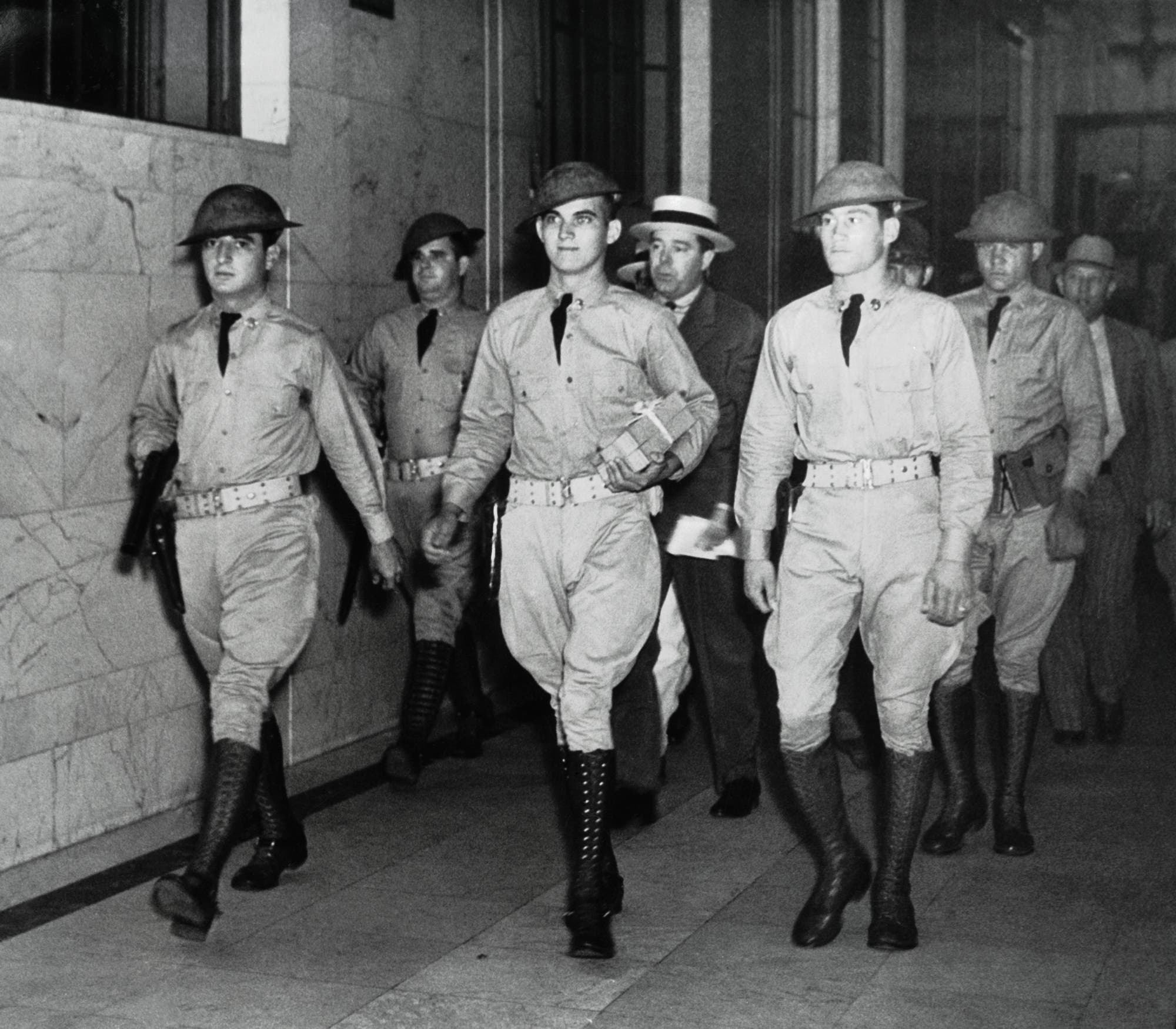 Eine Gruppe von uniformierten Männern in militärischer Kleidung marschiert durch einen Korridor in einem offiziellen Gebäude. Sie tragen Helme und Stiefel, einige halten Gegenstände in den Händen. Im Hintergrund sind weitere Personen zu sehen, darunter im Zentrum ein Mann mit Hut – Long. 