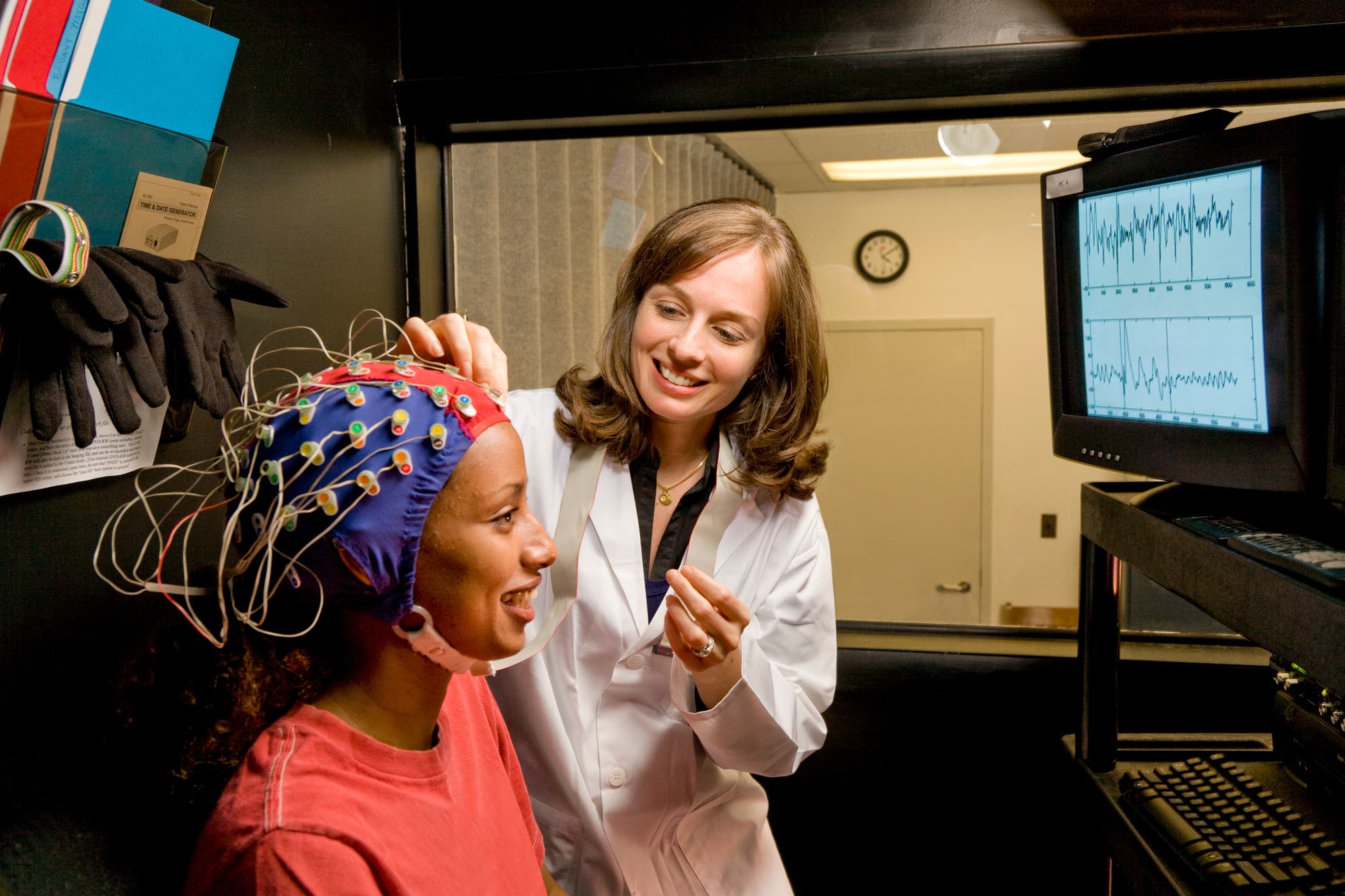 Eine Wissenschaftlerin in einem weißen Laborkittel befestigt Elektroden an einer EEG-Haube, die von einer sitzenden Person getragen wird. Neben ihnen zeigt ein Monitor EEG-Daten in Form von Wellenmustern. Die Szene findet in einem Labor statt, das mit wissenschaftlichen Geräten ausgestattet ist.
