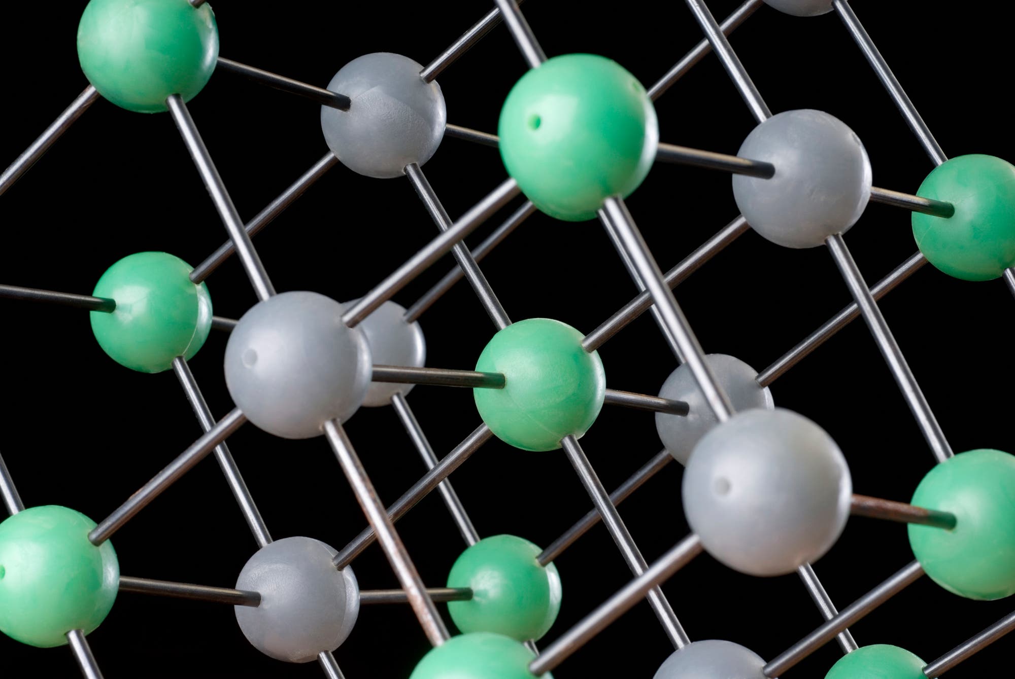 3D-Darstellung eines Kristallgitters mit grauen und grünen Kugeln, die Atome darstellen, verbunden durch silberne Stäbe, die chemische Bindungen symbolisieren. Die Struktur zeigt die regelmäßige Anordnung der Atome im Raum. Der Hintergrund ist schwarz, um den Fokus auf das Modell zu lenken.