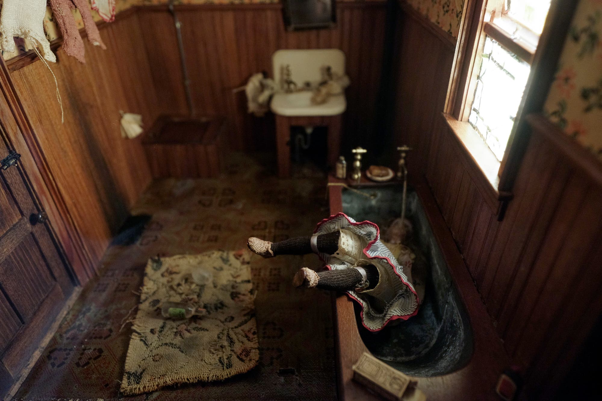 Ein altes Badezimmer mit Holzwänden und einem kleinen Fenster. In der Mitte steht eine Badewanne, in der eine Puppe mit einem rot-weißen Kleid kopfüber liegt, die Beine ragen unnatürlich heraus. Auf dem Boden liegt ein Teppich mit einer Schnapsflasche und einem Glas. Im Hintergrund befindet sich ein Waschbecken mit einigen Gegenständen. Die Szene wirkt düster und altmodisch.