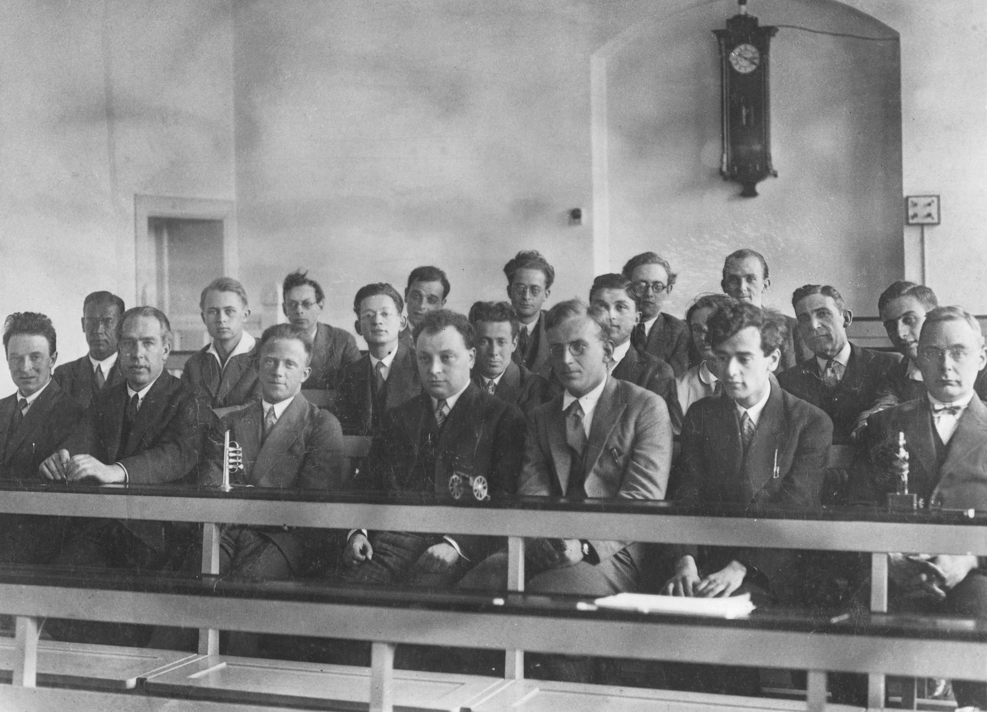 Eine Gruppe von Männern in Anzügen sitzt in einem Klassenzimmer oder Konferenzraum. Die mehrheitlich jungen Männer auf dem historischen Schwarz-weiß-Foto blicken aufmerksam nach vorne. Im Hintergrund hängt eine große Wanduhr. Die Atmosphäre wirkt formell und konzentriert. Der Raum ist schlicht eingerichtet mit weißen Wänden.