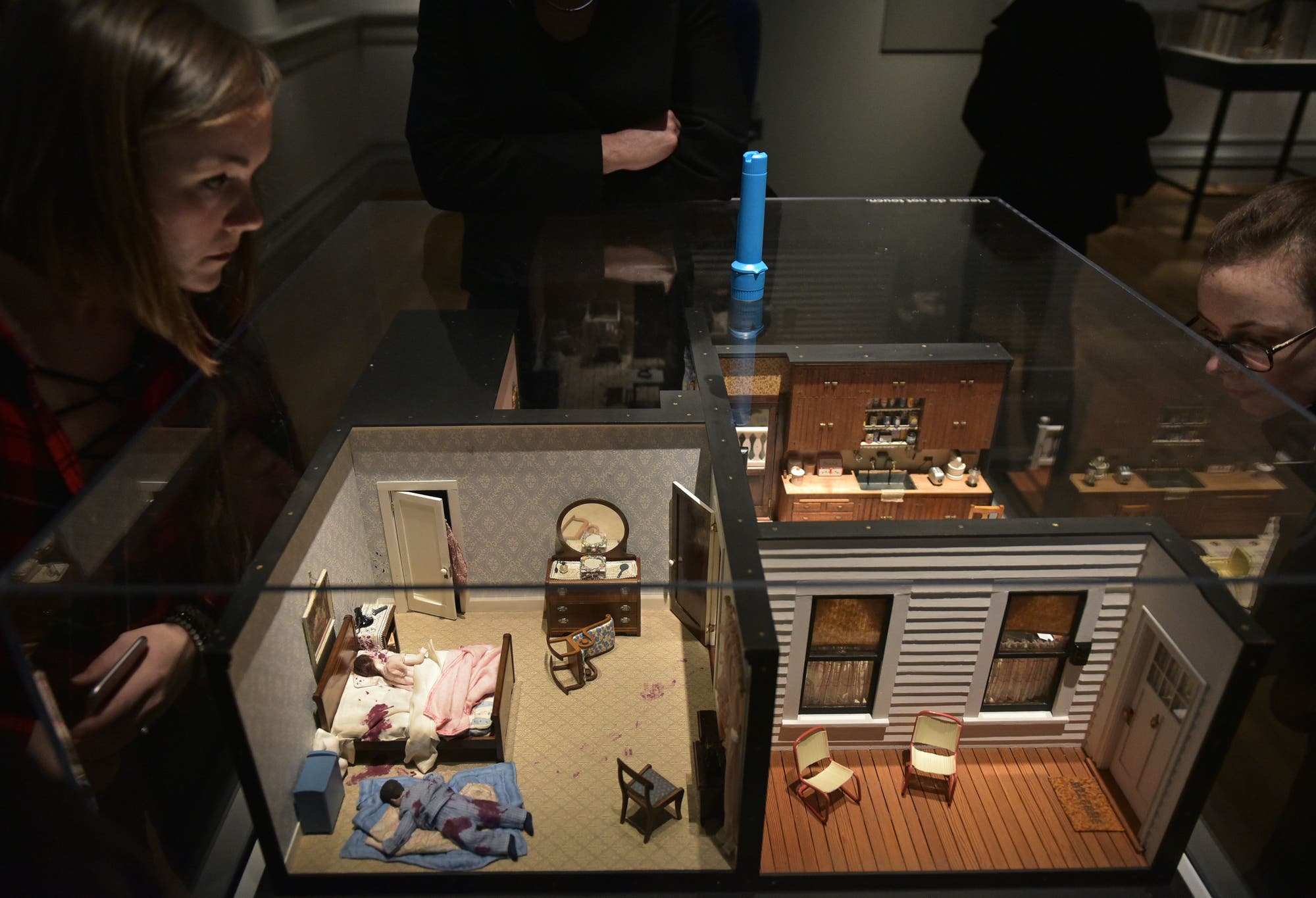 Eine Miniatur-Diorama-Szene in einem Museum, die ein Schlafzimmer und eine Küche in einem Puppenhaus zeigt. Zwei Personen betrachten das Diorama von oben, eine links und eine rechts. Das Schlafzimmer enthält ein Bett mit rosa Decke, einen Schminktisch mit Spiegel und einen Stuhl. Die Küche ist mit Schränken und Haushaltsgegenständen ausgestattet. Die Szene ist in einem Glaskasten ausgestellt.