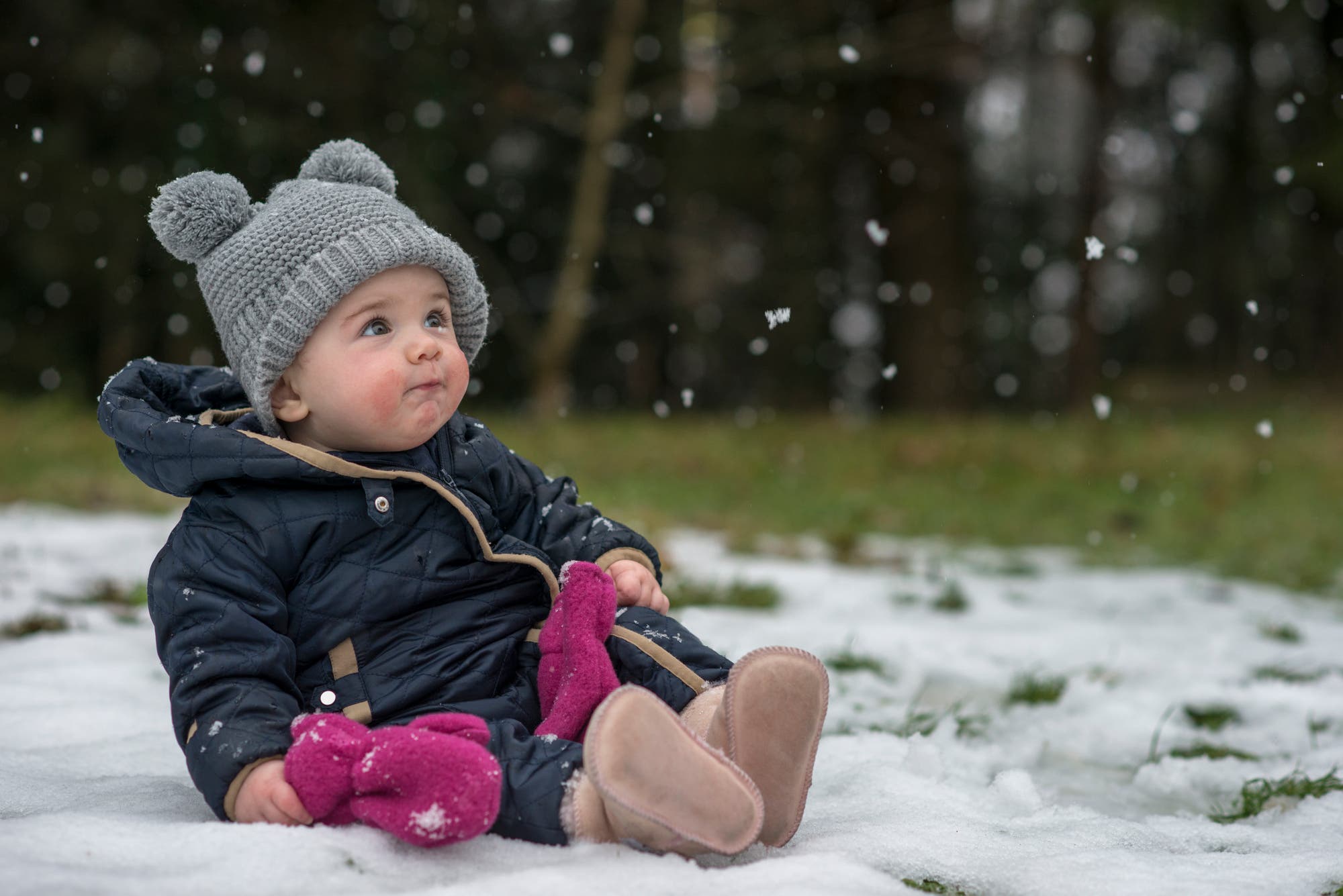 Ein kleines Kind sitzt in einem schneebedeckten Park und schaut nach oben, während Schneeflocken um es herum fallen. Es trägt eine graue Mütze mit Bommeln, einen dunkelblauen Schneeanzug und hält pinke Handschuhe. Der Hintergrund zeigt unscharfe Bäume.