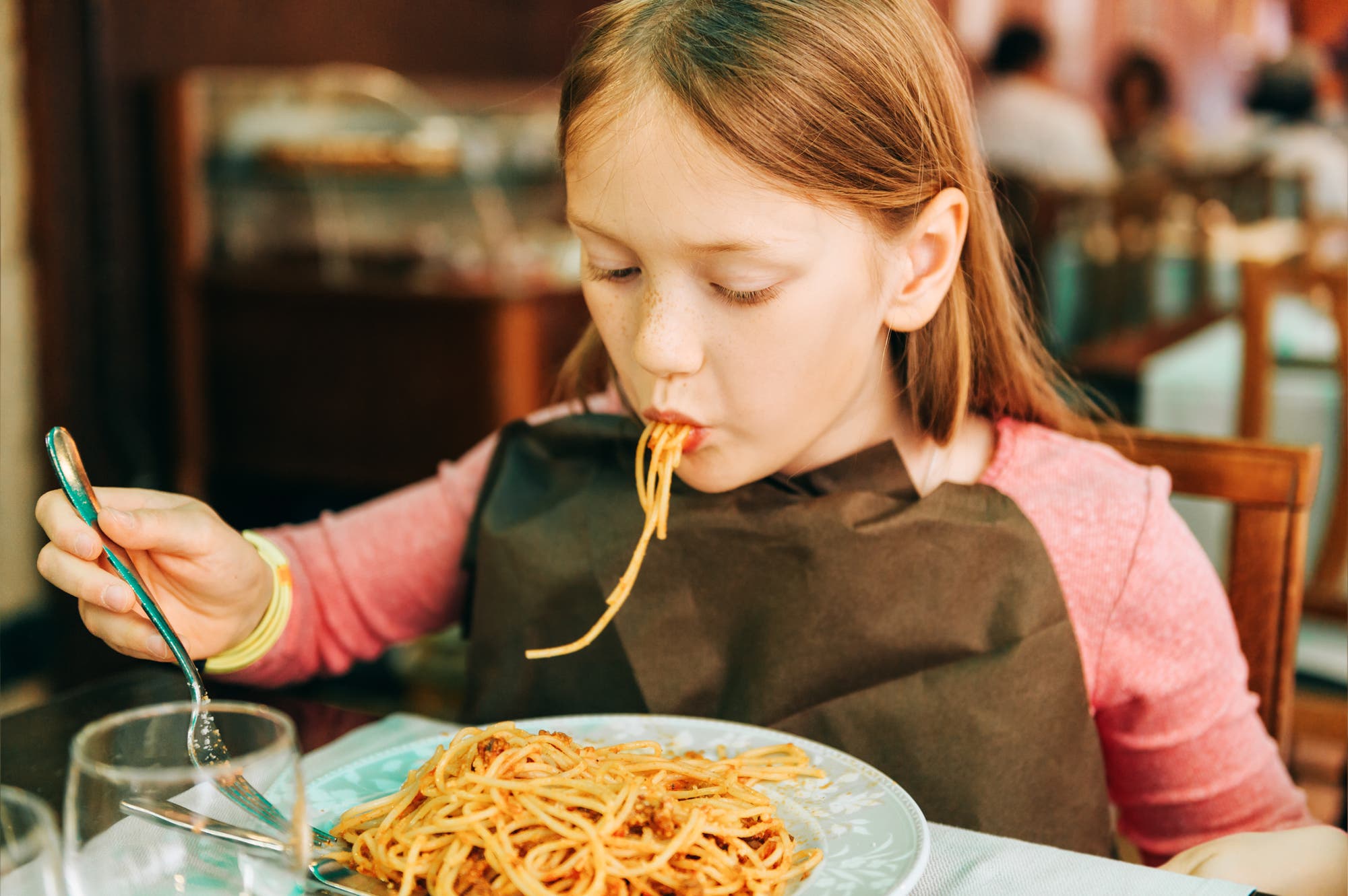 Ein rothaariges Mädchen isst in einem Restaurant Spaghetti; ein paar Nudeln hängen noch zur Hälfte aus seinem Mund heraus. Es hält eine Gabel in der Hand; auf dem Tisch vor ihm steht ein voller Teller mit Spaghetti bolognese. Im Hintergrund sieht man weitere Leute an Tischen sitzen.