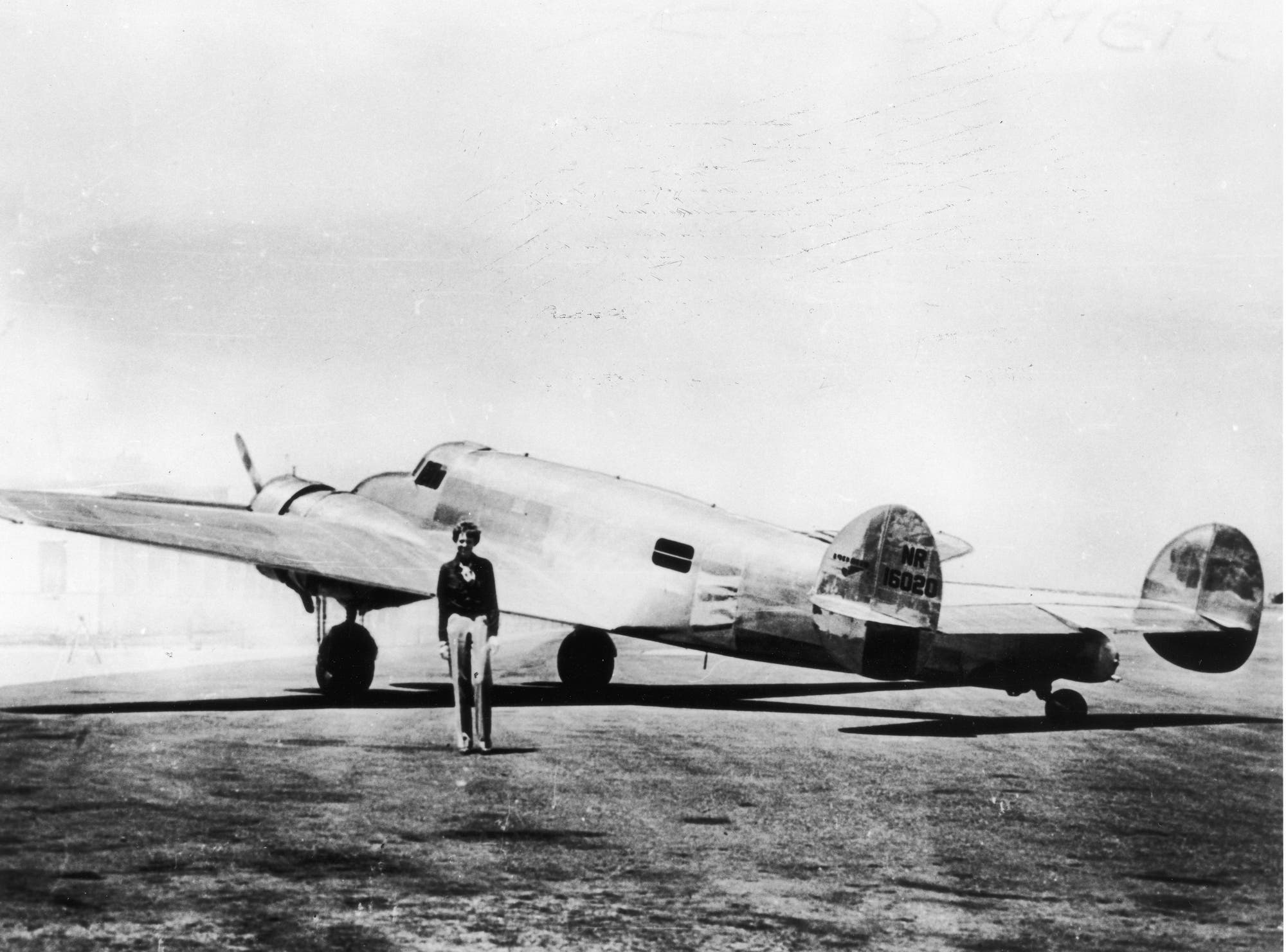 Amelia Earhart steht vor einem zweimotorigen Flugzeug auf einem Flugfeld. Das Flugzeug hat eine glänzende Metalloberfläche und trägt die Kennzeichnung "NR16020" am Heck. Der Himmel ist klar und der Boden asphaltiert. Earhart trägt eine dunkle Jacke und helle Hosen.