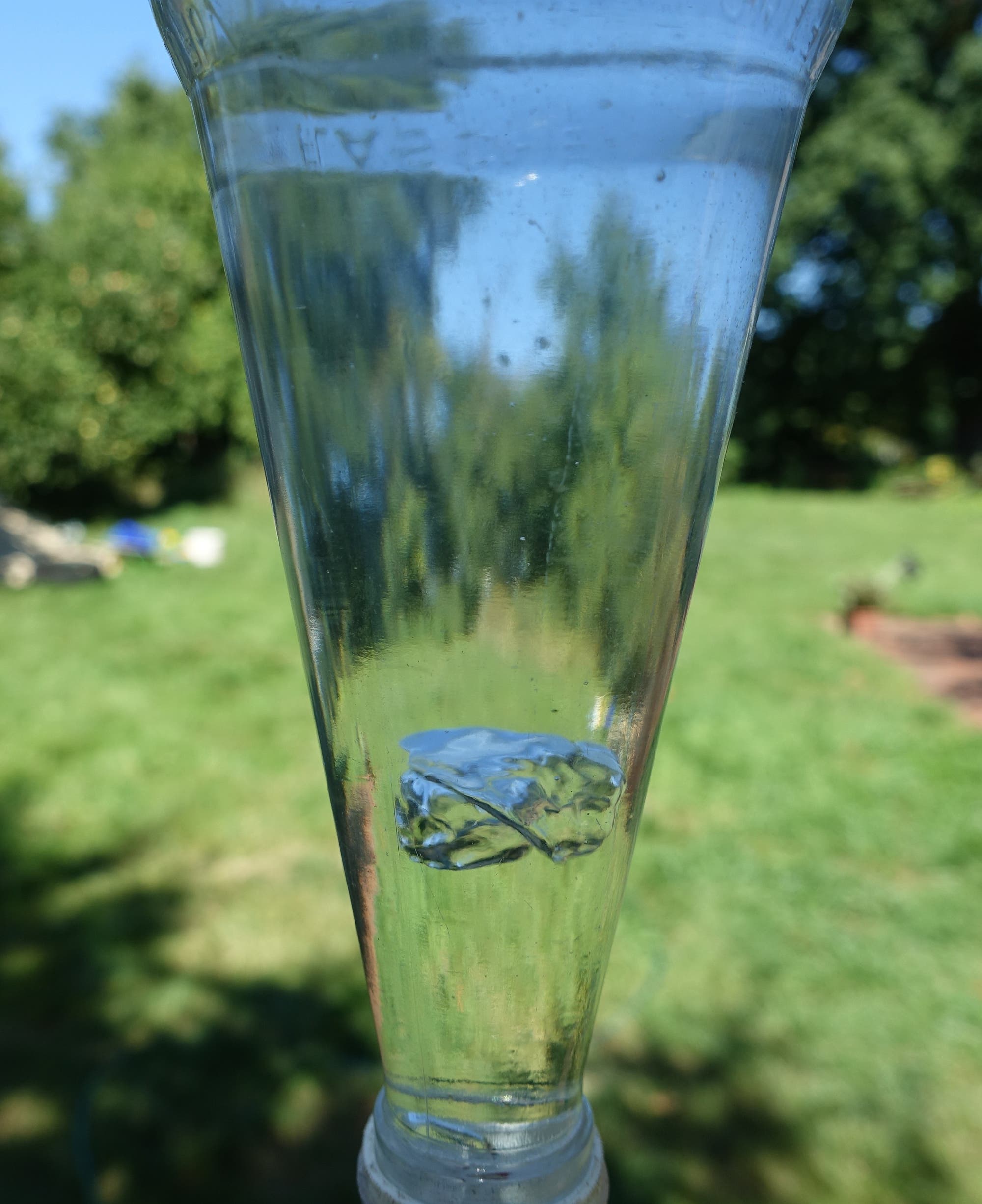 Eine Glasflasche wird kopfüber vor einem grünen, unscharfen Gartenhintergrund gehalten. Im Inneren des Gefäßes befindet sich eine schwebende Luftblase.