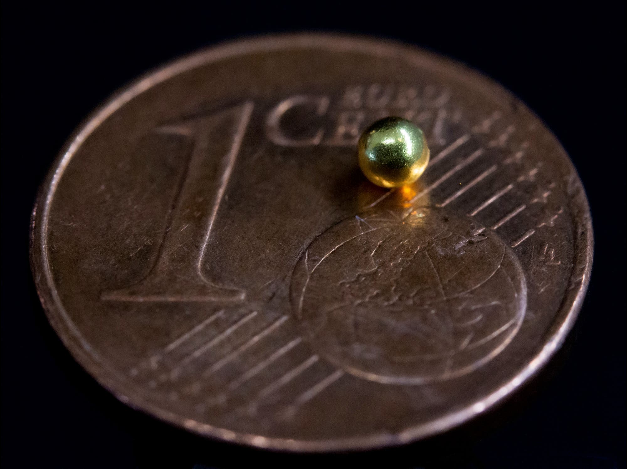 Eine kleine, goldene Kugel liegt auf einer 1-Cent-Euro-Münze. Die Münze zeigt die Zahl "1" und das Wort "Euro Cent" sowie ein stilisiertes Globus-Design. Die Kugel hebt sich durch ihre glänzende Oberfläche von der matten Münze ab und verdeutlicht den Größenvergleich zwischen der Kugel und der Münze.