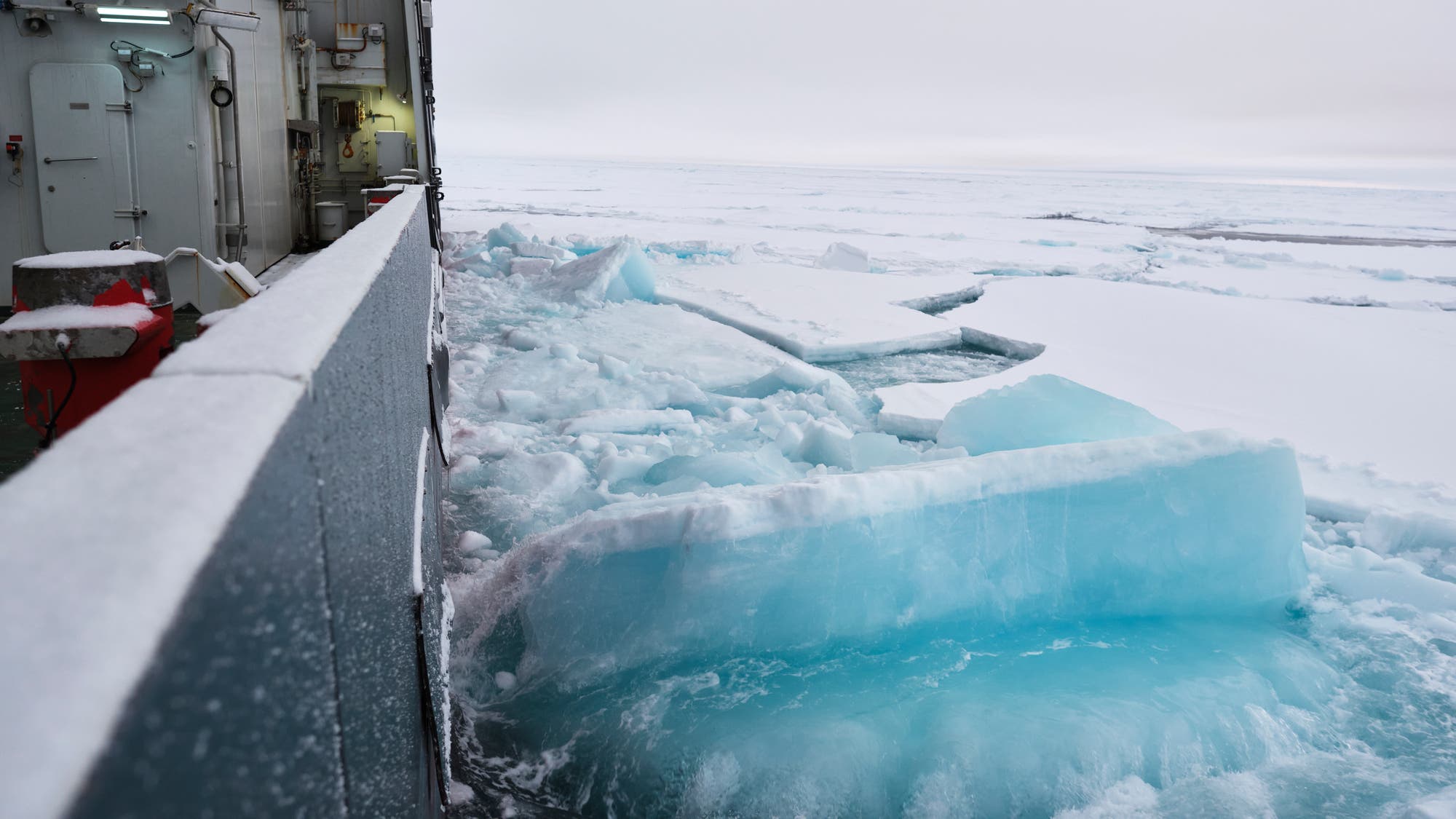 Ein Schiff bricht durch dickes Meereis in einer arktischen Umgebung. Die Eisschollen sind blau und weiß gefärbt, und das Wasser ist teilweise sichtbar. Die Bordwand des Schiffes ist links im Bild zu sehen, während sich der Horizont in der Ferne erstreckt. Die Szene vermittelt eine kalte, isolierte Atmosphäre.