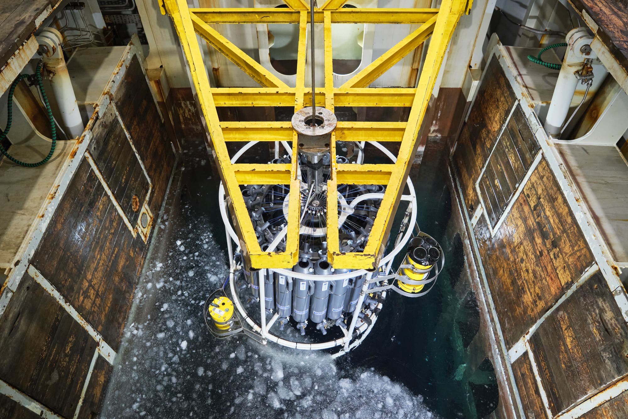 Ein gelber Kran hebt ein wissenschaftliches Gerät, das aus mehreren zylindrischen Behältern besteht, aus einem eisbedeckten Gewässer. Das Gerät befindet sich in einem Schacht auf einem Schiff. Die Szene zeigt den Einsatz von Meeresforschungstechnologie zur Probenentnahme.