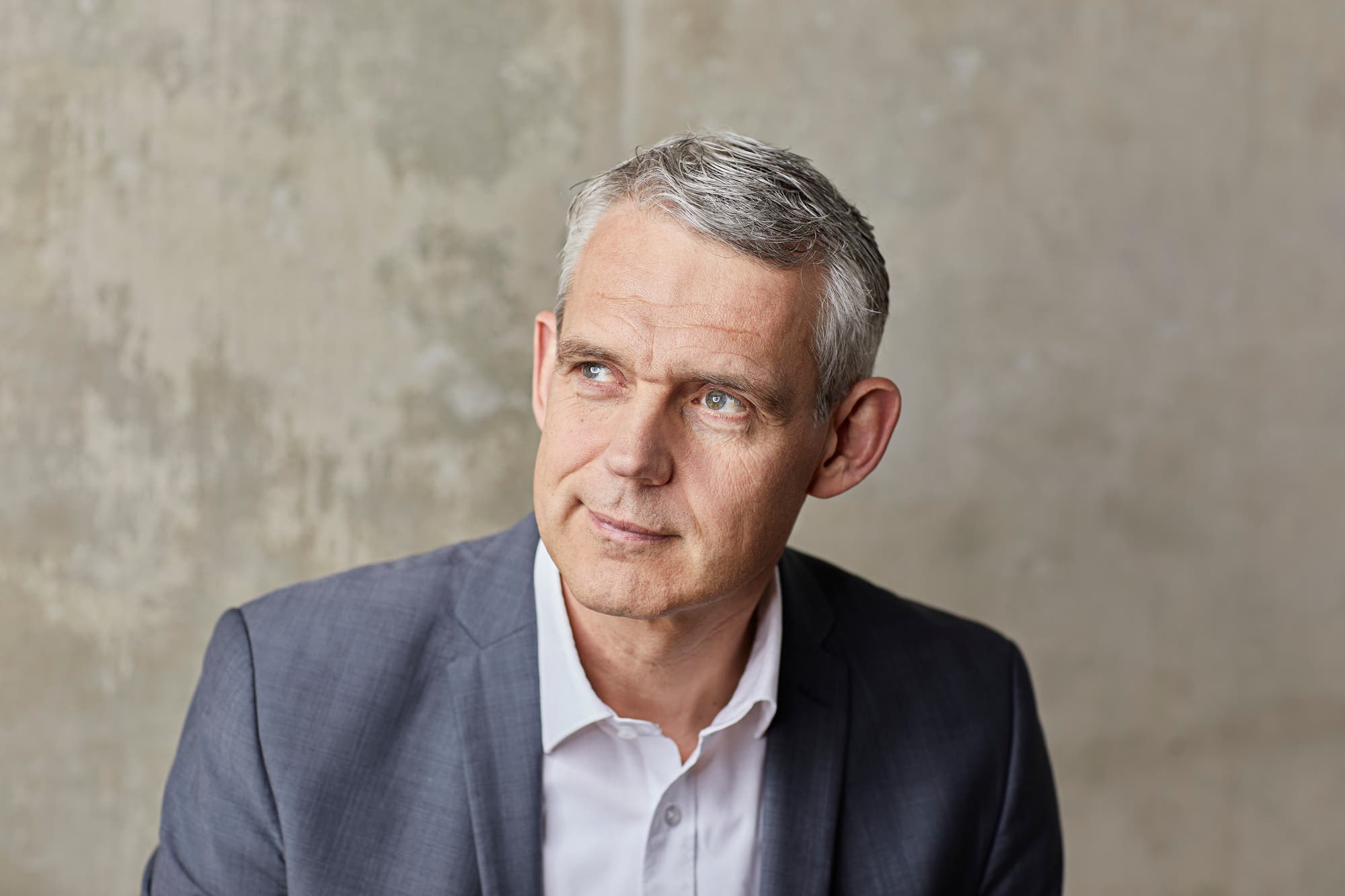 Eine Person in einem grauen Anzug und weißem Hemd blickt nachdenklich zur Seite. Der Hintergrund ist eine schlichte, strukturierte Wand. Die Szene vermittelt einen professionellen und reflektierten Eindruck.