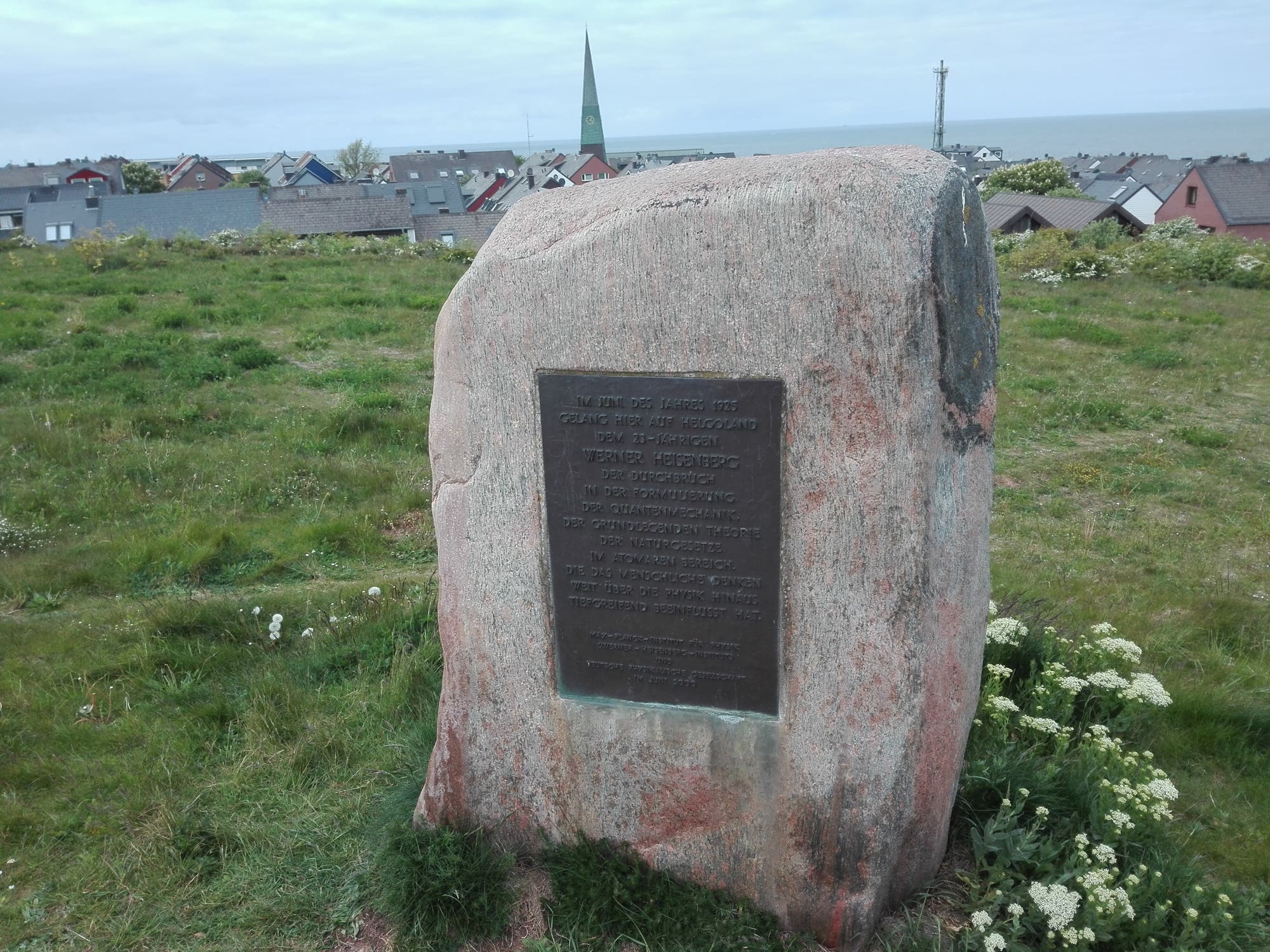 Ein Gedenkstein mit einer Inschrift steht auf einer grünen Wiese. Im Hintergrund sind Häuser mit grauen Dächern und eine Kirche mit einem spitzen Turm zu sehen. Der Himmel ist bewölkt, und im Hintergrund erstreckt sich das Meer.