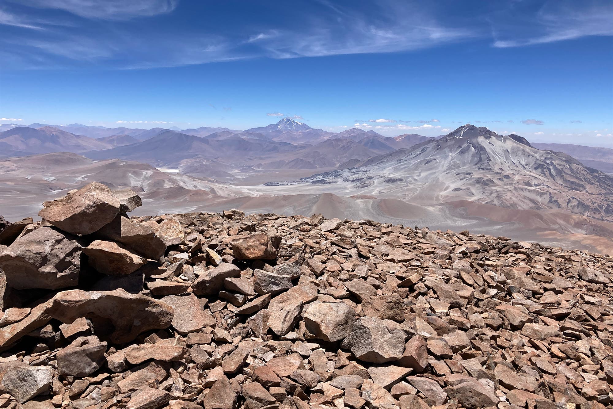Vista panorámica de los picos volcánicos de Atacama: en primer plano hay un guijarro marrón, al fondo se pueden ver algunos volcanes, el cielo es azul, el resto tiene diferentes tonalidades de marrón.
