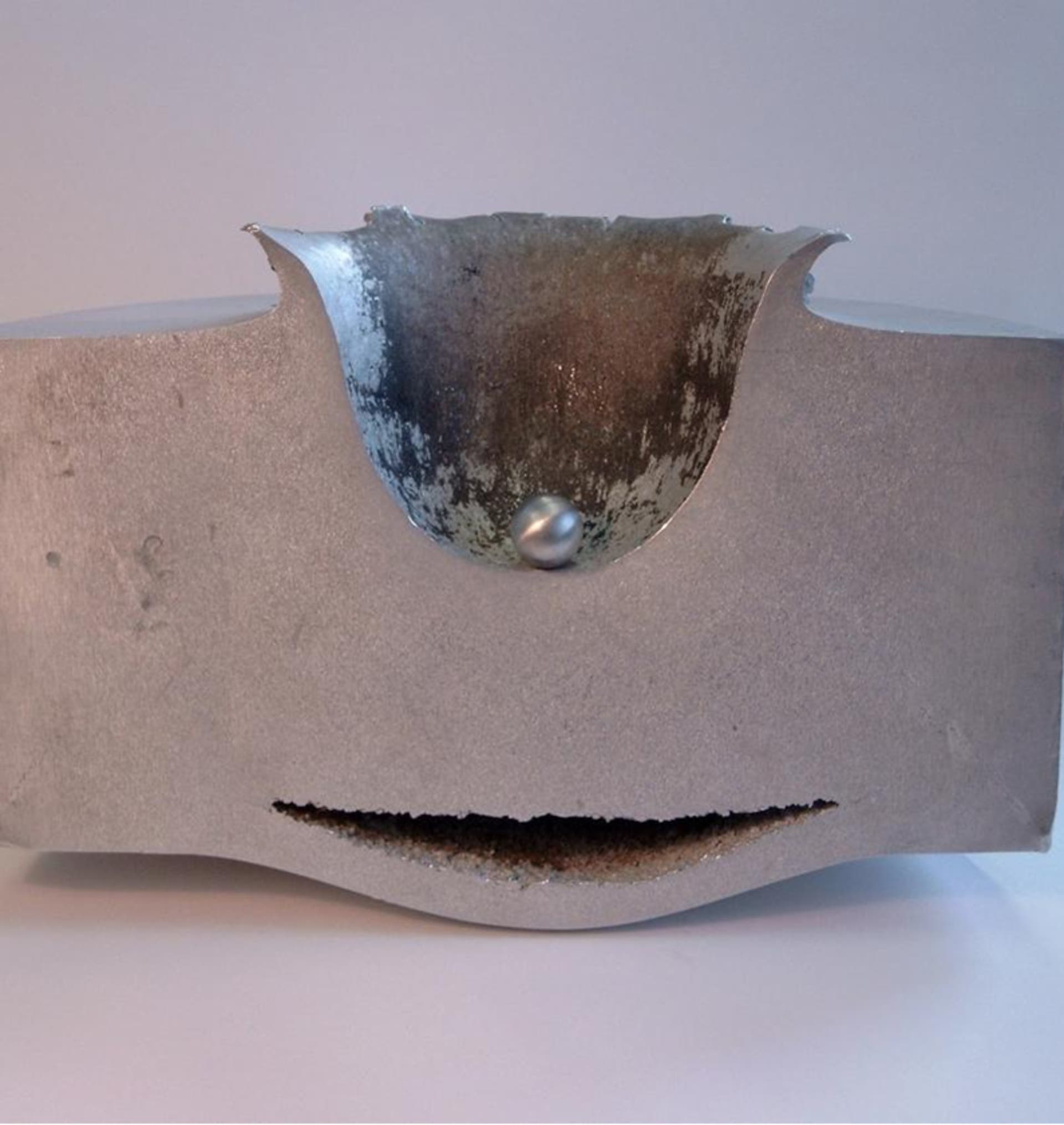 Ein Metallblock mit einer deutlichen Verformung und einem Riss, der durch den Aufprall eines Projektils verursacht wurde. Das Projektil steckt teilweise in der Oberseite des Blocks, umgeben von einer aufgewölbten und beschädigten Metallfläche. Der Hintergrund ist neutral und lenkt nicht vom Hauptmotiv ab.