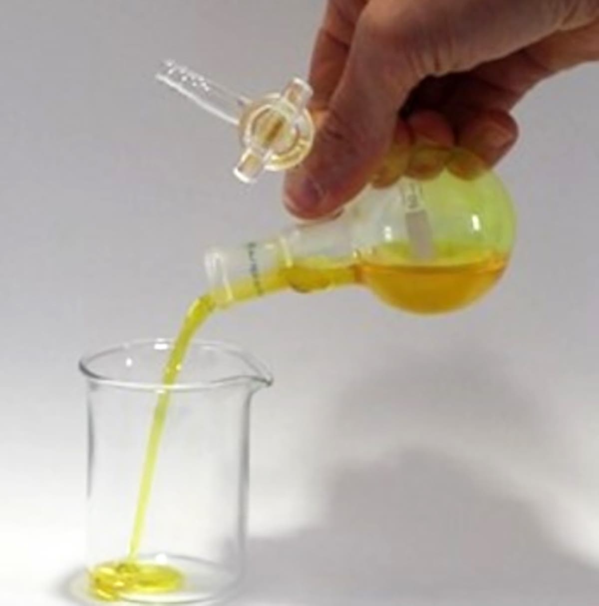 Eine Hand hält eine runde Glasflasche mit gelber Flüssigkeit, die in ein Becherglas gegossen wird. Die Flüssigkeit fließt in einem dünnen Strahl in das Becherglas. Die Szene findet auf einem neutralen Hintergrund statt.