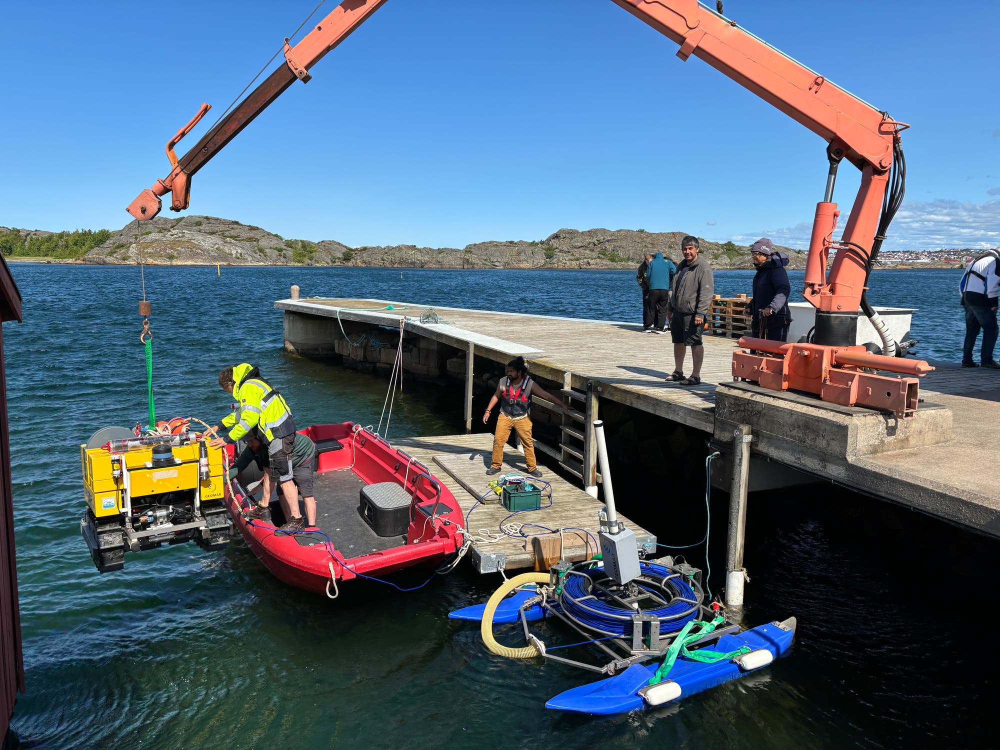 Ein rotes Schlauchboot liegt an einem Steg, während ein Kran ein gelbes Gerät ins Wasser hebt. Zwei Personen auf dem Boot arbeiten mit dem Gerät, während eine weitere Person auf dem Steg steht. Im Hintergrund sind felsige Inseln und ein klarer Himmel zu sehen. Der Kran ist orange und auf dem Steg befestigt. Die Szene zeigt eine maritime Arbeitsumgebung.