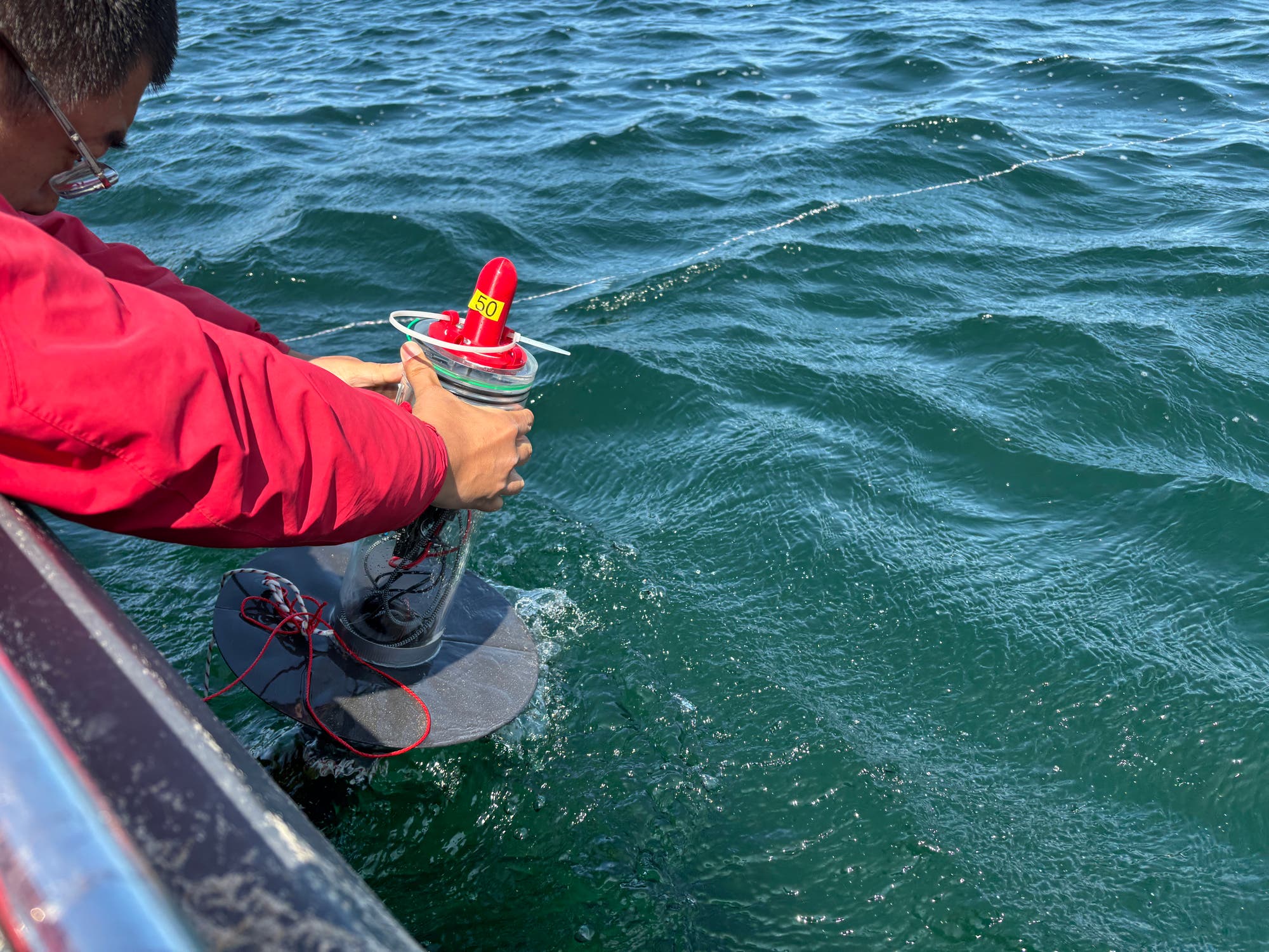 Eine Person in roter Jacke hält ein wissenschaftliches Gerät über die Seite eines Bootes, das teilweise im Wasser ist. Das Gerät hat eine rote Spitze mit der Aufschrift "360". Die Szene spielt sich auf einem Gewässer mit leichtem Wellengang ab.