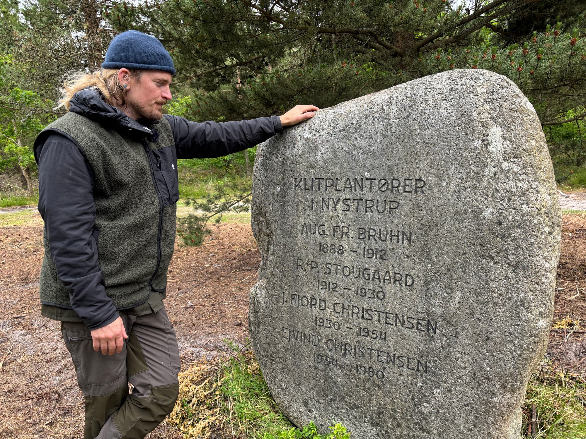 Eine Person steht neben einem großen Gedenkstein in einem bewaldeten Gebiet. Der Stein trägt die Inschrift „KLITPLANTØRER I NYSTRUP“ gefolgt von mehreren Namen und Jahreszahlen, darunter „AUG. FR. BRUHN 1888-1912“ und „P. P. STOUGAARD 1912-1930“. Die Person trägt eine Mütze und eine Outdoor-Jacke und berührt den Stein mit einer Hand. Im Hintergrund sind Bäume und Sträucher zu sehen.