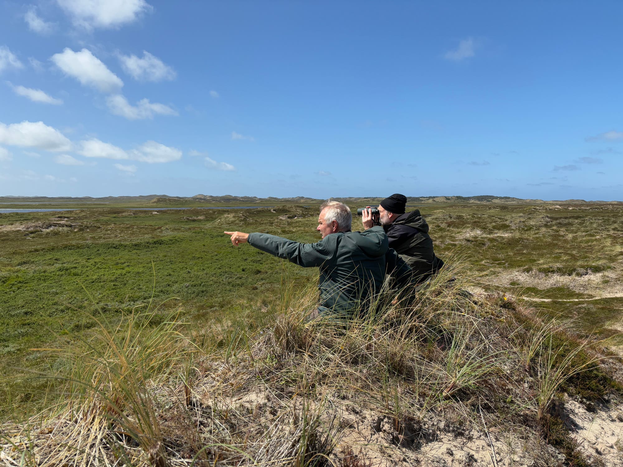 Zwei Personen stehen auf einer grasbewachsenen Düne und blicken in die Ferne. Eine Person zeigt mit dem Finger in die Landschaft, während die andere durch ein Fernglas schaut. Der Himmel ist klar mit einigen Wolken, und die Umgebung besteht aus weitläufigen, grünen Hügeln und Dünen.
