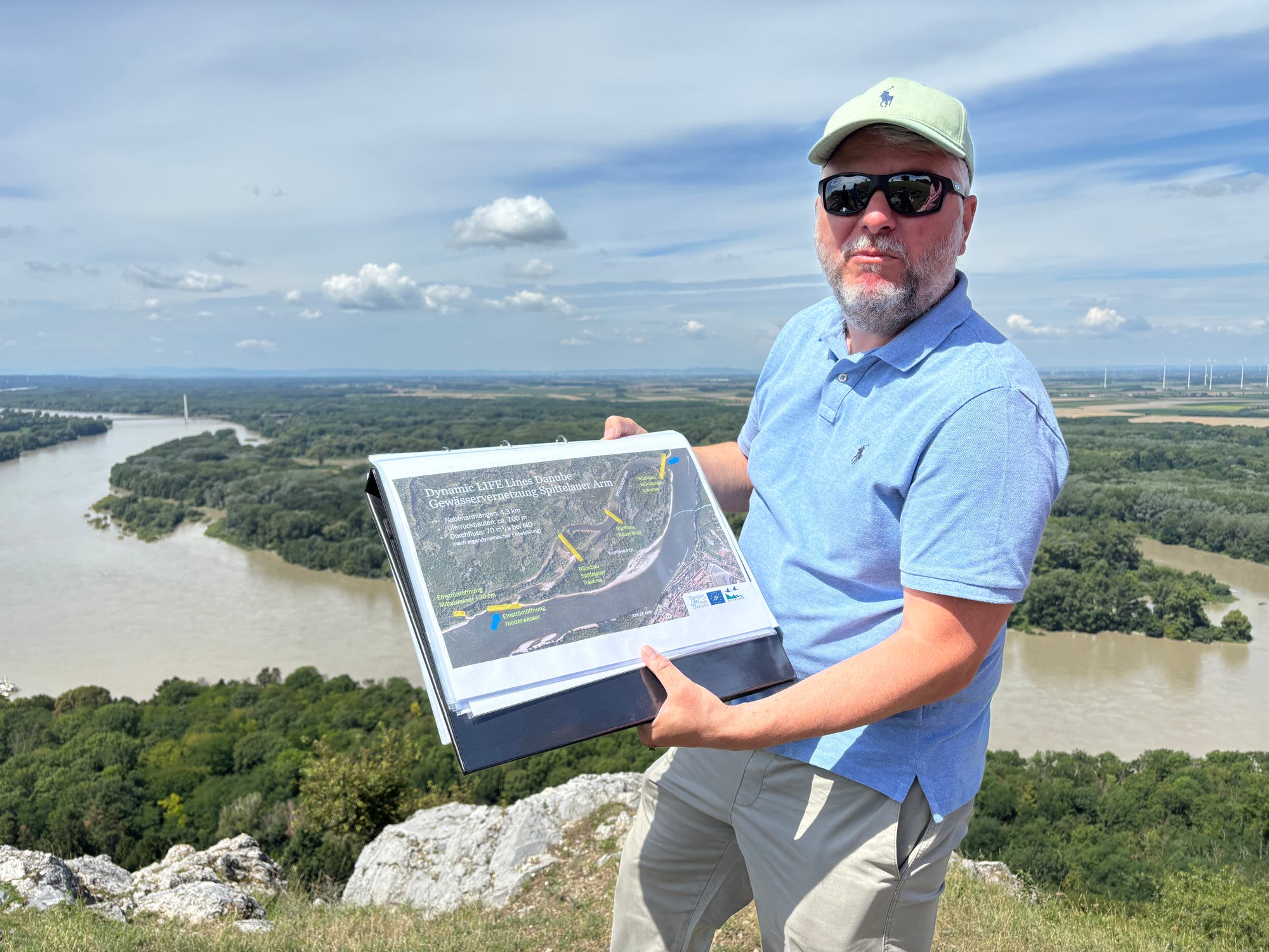 Eine Person steht auf einem Hügel mit Blick auf einen Fluss und hält ein Informationsblatt mit einer Karte und Text. Die Landschaft im Hintergrund zeigt einen breiten Fluss, Wälder und Windräder am Horizont. Der Himmel ist leicht bewölkt. Die Person trägt eine Sonnenbrille, einen Hut und ein hellblaues Hemd.