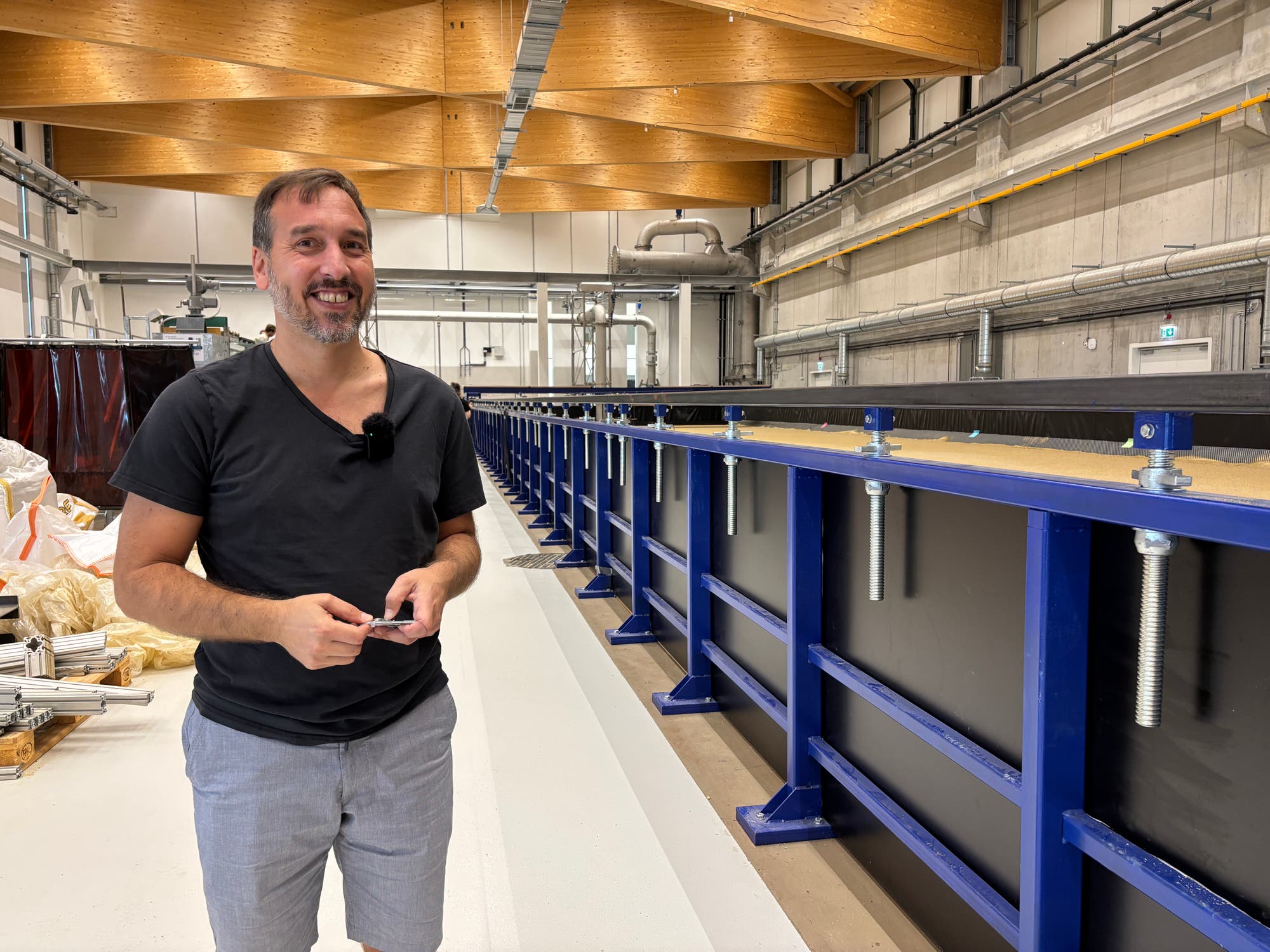 Eine Person steht in einer großen, modernen Industriehalle neben einer langen, blauen Metallstruktur, die Teil einer wissenschaftlichen Anlage ist. Die Halle hat eine hohe Decke mit sichtbaren Holzbalken und zahlreichen Rohren und Leitungen an den Wänden. Die Person trägt ein Mikrofon und lächelt in die Kamera. Im Hintergrund sind verschiedene technische Geräte und Materialien zu sehen.