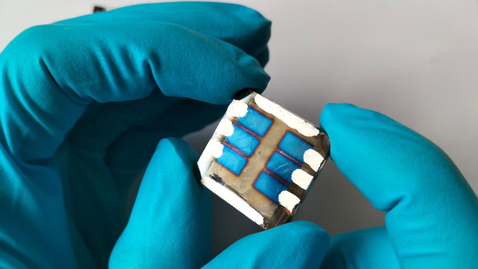 Eine Person mit blauen Handschuhen hält ein kleines, quadratisches elektronisches Bauteil. Das Bauteil hat eine blaue Oberfläche mit weißen Linien und Kontakten. Der Hintergrund ist neutral und hell.