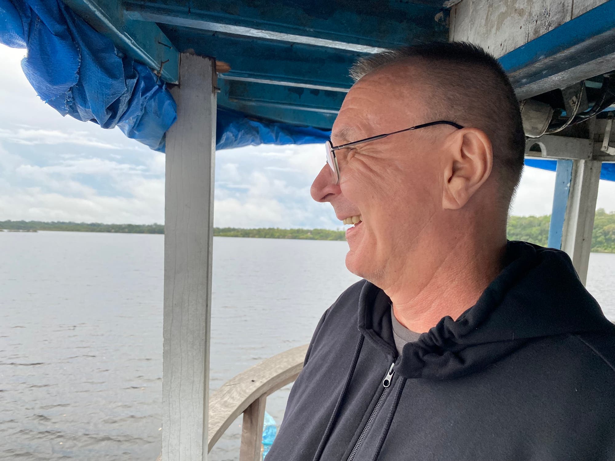 Ein Mann steht auf einem Boot und blickt lächelnd über das Wasser, im Hintergrund sieht man Wolken am Himmel und ein bewaldetes Ufer. Er trägt eine Brille und eine dunkle Jacke. Die Szene vermittelt den Eindruck von Ruhe und Freude.