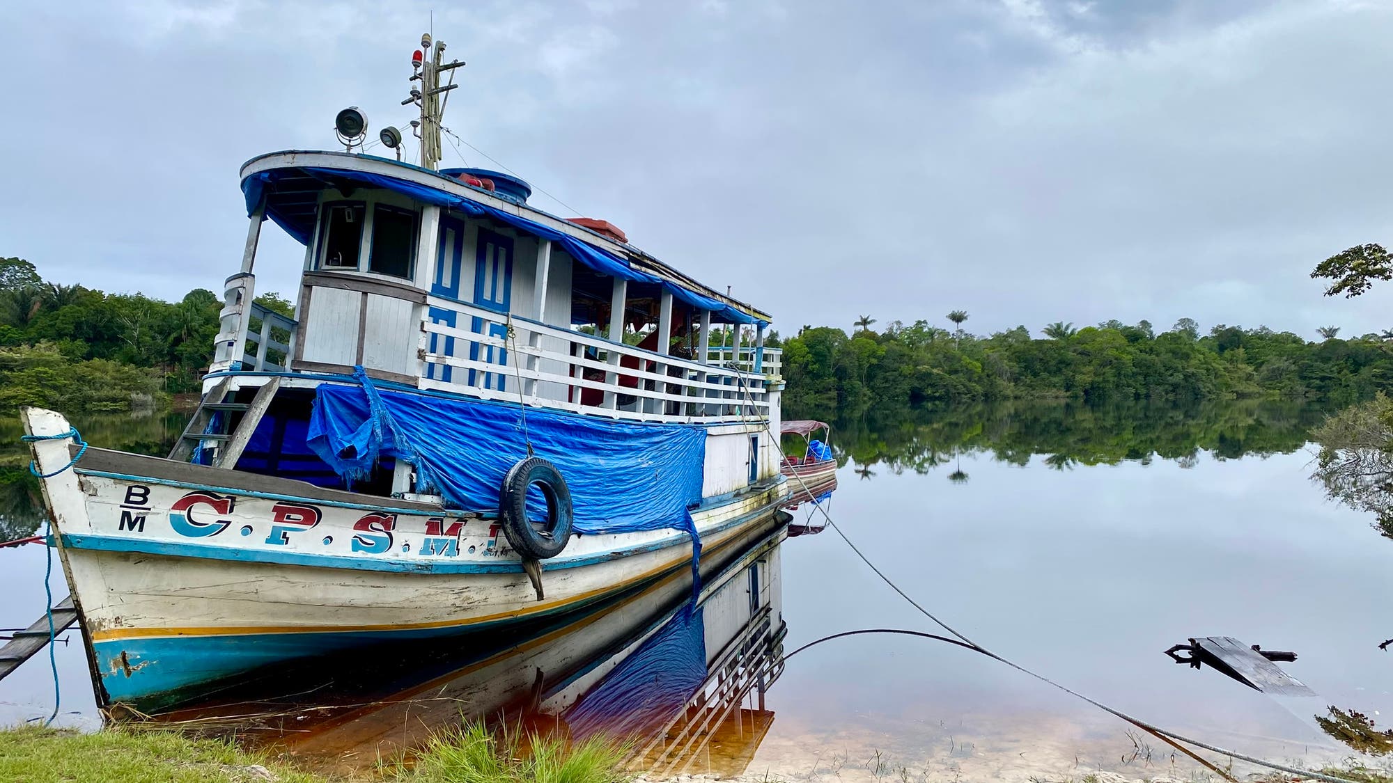 Ein traditionelles Holzboot mit blauer und weißer Bemalung liegt an einem ruhigen Flussufer vor Anker. Das Boot ist mit blauen Planen abgedeckt und trägt die Aufschrift "CPSML" am Bug. Im Hintergrund erstreckt sich ein dichter grüner Wald unter einem bewölkten Himmel. Das Wasser spiegelt die Landschaft und das Boot wider, was eine friedliche und idyllische Szene schafft.