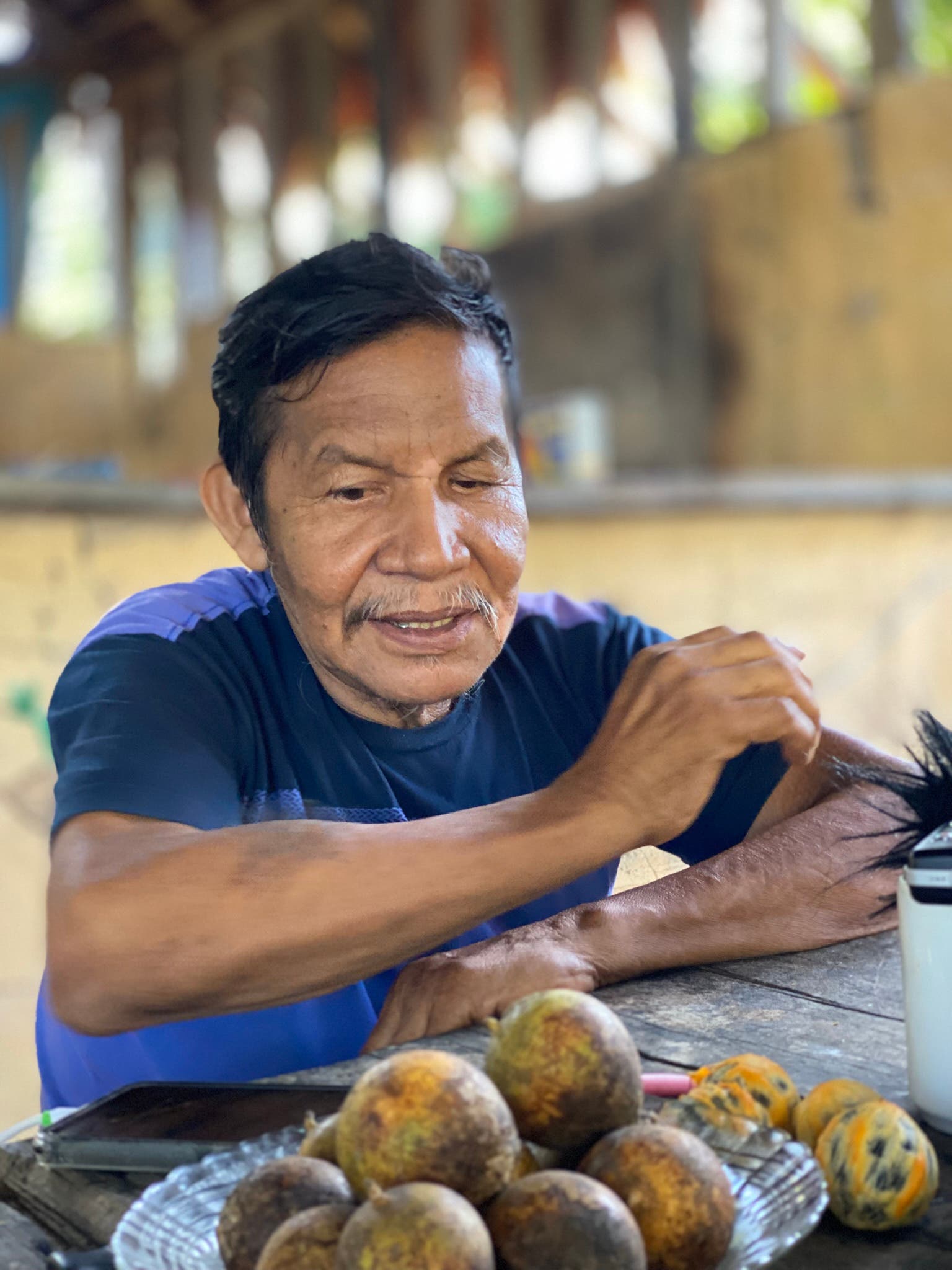 Ein Mann sitzt an einem Holztisch in einem rustikalen Raum. Vor ihm liegt ein Teller mit mehreren runden Früchten. Der Mann trägt ein dunkelblaues T-Shirt und scheint entspannt zu sein. Der Hintergrund ist unscharf, was den Fokus auf den Mann und die Früchte lenkt. Die Szene wirkt ruhig und alltäglich.