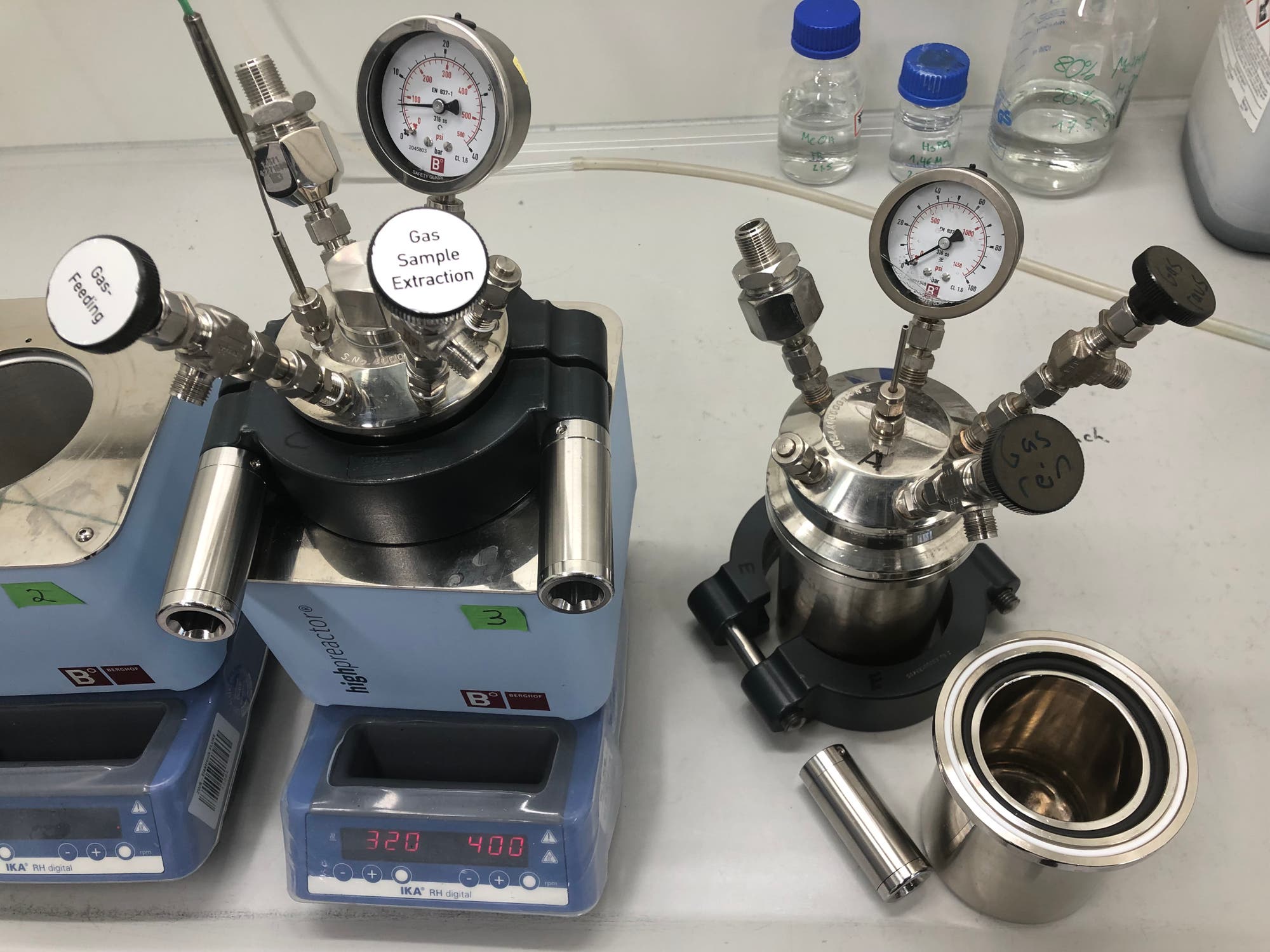 Zwei Laborreaktoren mit Druckanzeigen und Ventilen auf einem Tisch. Der linke Reaktor zeigt die Beschriftung "Gas Sample Extraction" und "Gas Feeding". Die digitale Anzeige zeigt "320" und "400". Im Hintergrund sind mehrere Flaschen mit blauen Verschlüssen zu sehen.