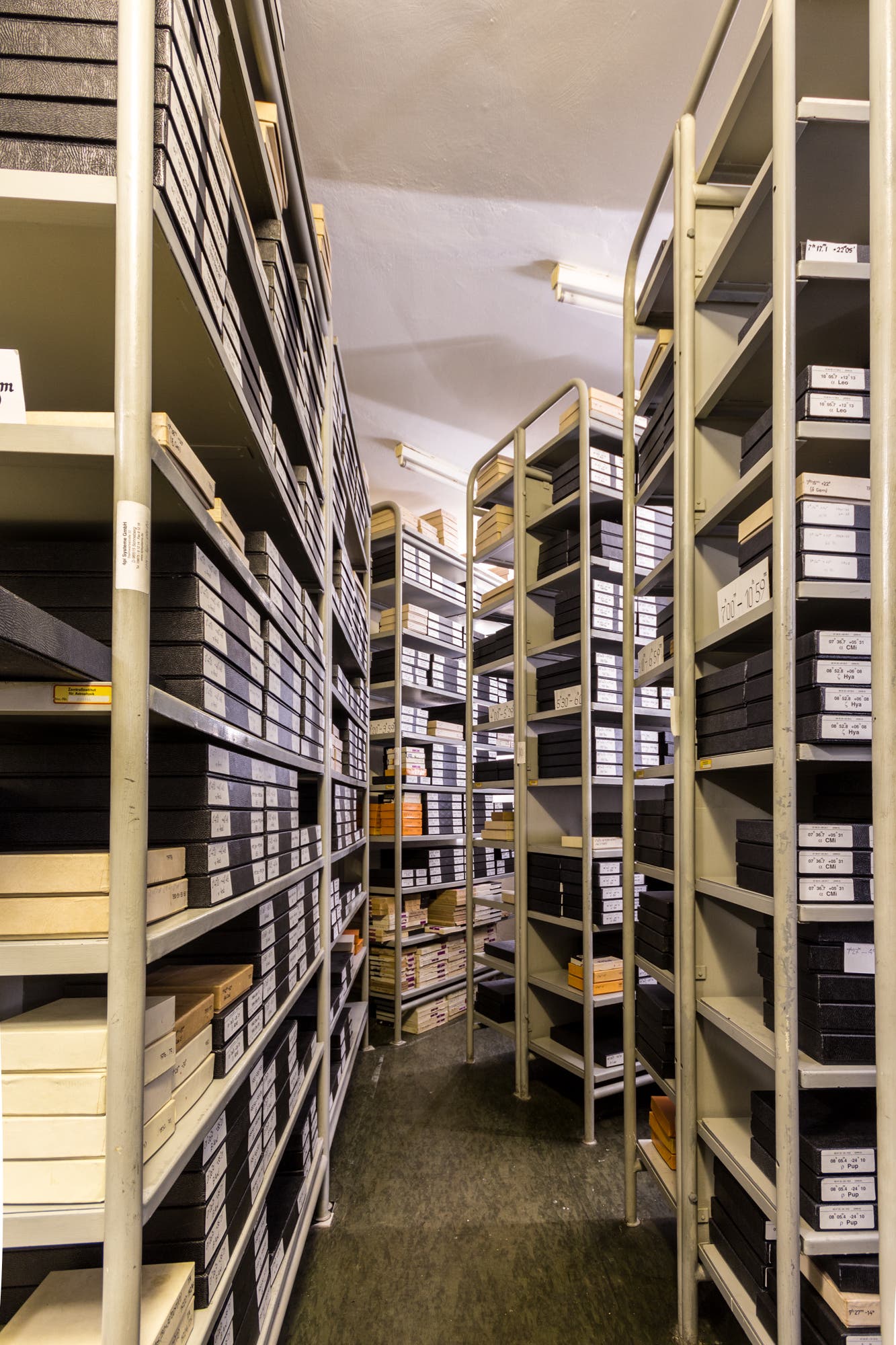 Ein Archivraum mit hohen offenen Regalen, die mit zahlreichen schwarzen und weißen Archivboxen gefüllt sind. Die Boxen sind ordentlich gestapelt und mit Etiketten versehen. Der Raum ist gut beleuchtet und die Regale sind aus Metall. Der Boden ist mit einem dunkelgrauen Teppich ausgelegt.