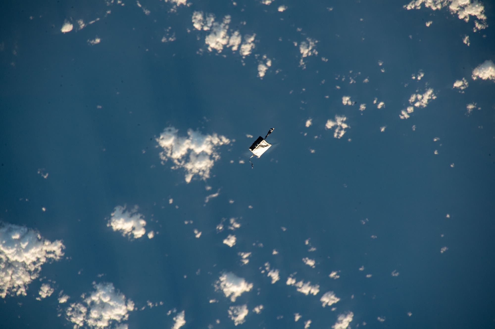 Eine Werkzeugtasche in Weiß fliegt durch das All und überquert einen blauen Ozean mit Wolken am Himmel.