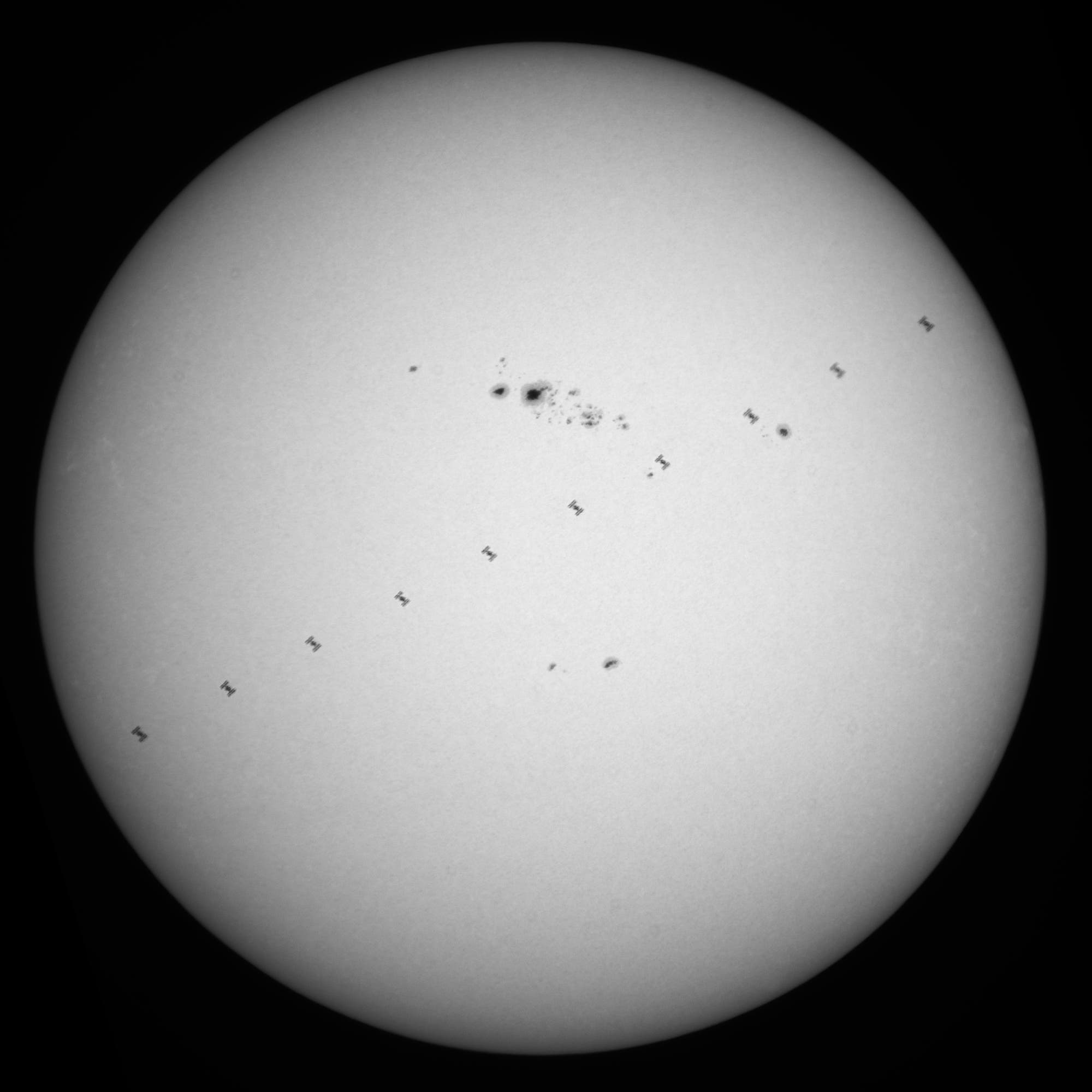 La Estación Espacial Internacional (ISS) pasa frente al sol, visible como una pequeña siluja en una línea diagonal. Se pueden ver varias manchas solares en la superficie del sol. La imagen muestra el contraste entre el sol brillante y el lado oscuro de la ISS.