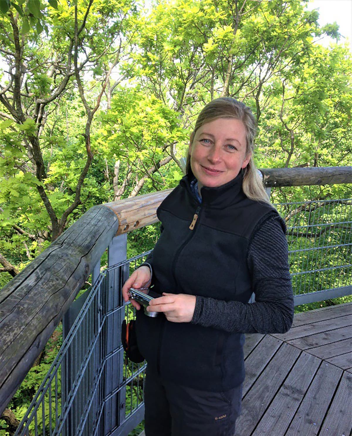 Eine Frau steht lächelnd auf einem hölzernen Aussichtsturm, umgeben von üppigem grünem Laub. Sie trägt eine dunkle Weste und hält ein Smartphone in der Hand. Die Szene vermittelt einen Eindruck von Natur und Entspannung.