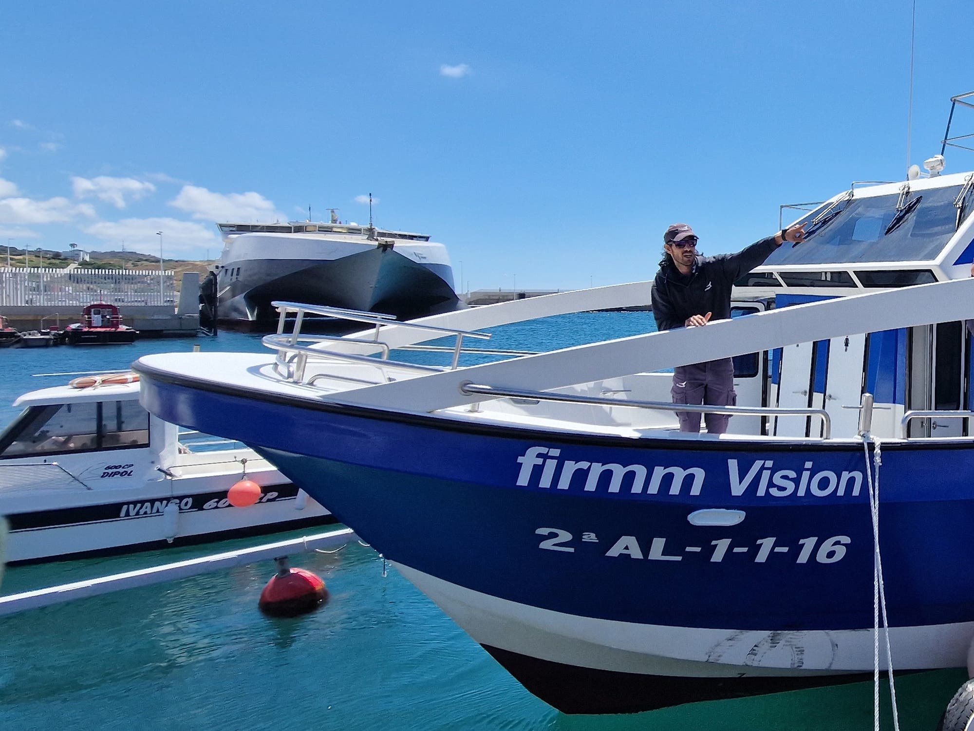 Ein Mann steht auf dem Deck eines blauen Bootes mit der Aufschrift "firmm Vision" und der Kennung "2ª AL-1-1-16". Er zeigt auf etwas außerhalb des Bildes. Im Hintergrund sind ein weiteres Boot und eine große Fähre im Hafen zu sehen. Der Himmel ist klar und blau.