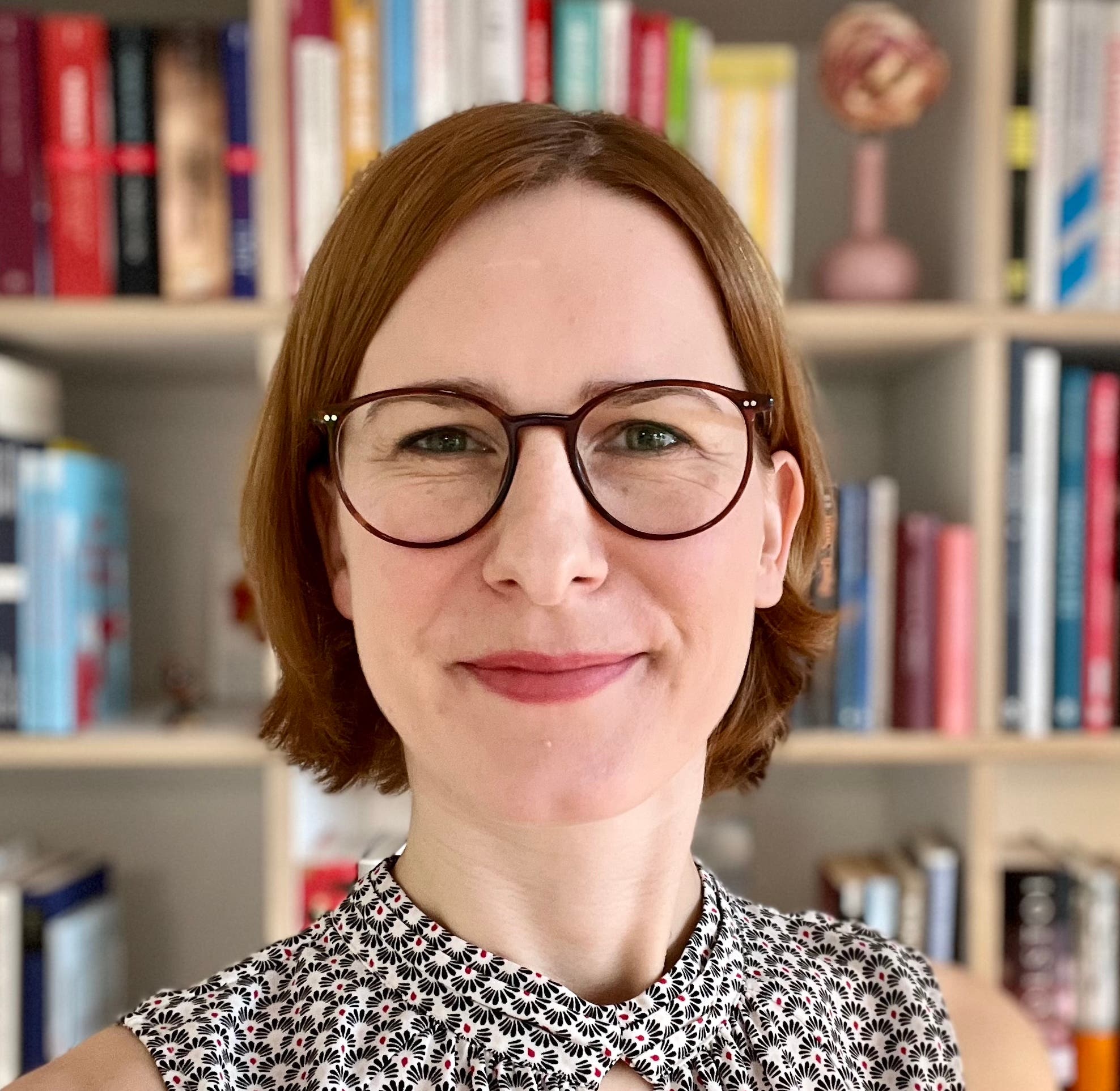Eine Frau mit Brille und kurzem, braunem Haar lächelt in die Kamera. Sie trägt ein ärmelloses, gemustertes Oberteil. Im Hintergrund ist ein Bücherregal mit vielen Büchern unscharf zu sehen.