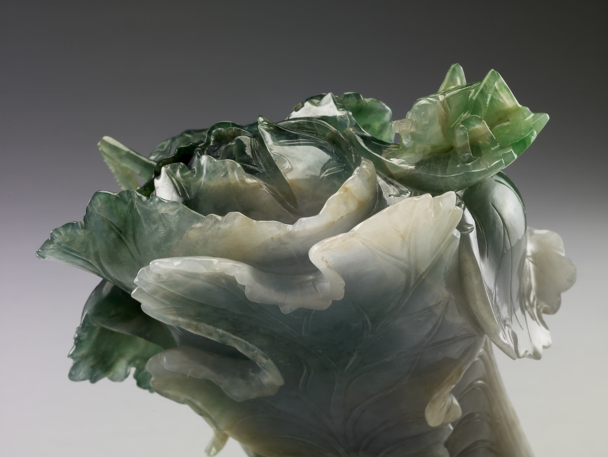 Eine kunstvolle Skulptur aus Jade, die einen Kohlkopf darstellt. Die Skulptur zeigt detaillierte, geschwungene Blätter in verschiedenen Grüntönen, die die natürliche Textur und Form eines echten Kohls nachahmen. Die Oberfläche ist glatt und glänzend, was die feine Handwerkskunst betont.