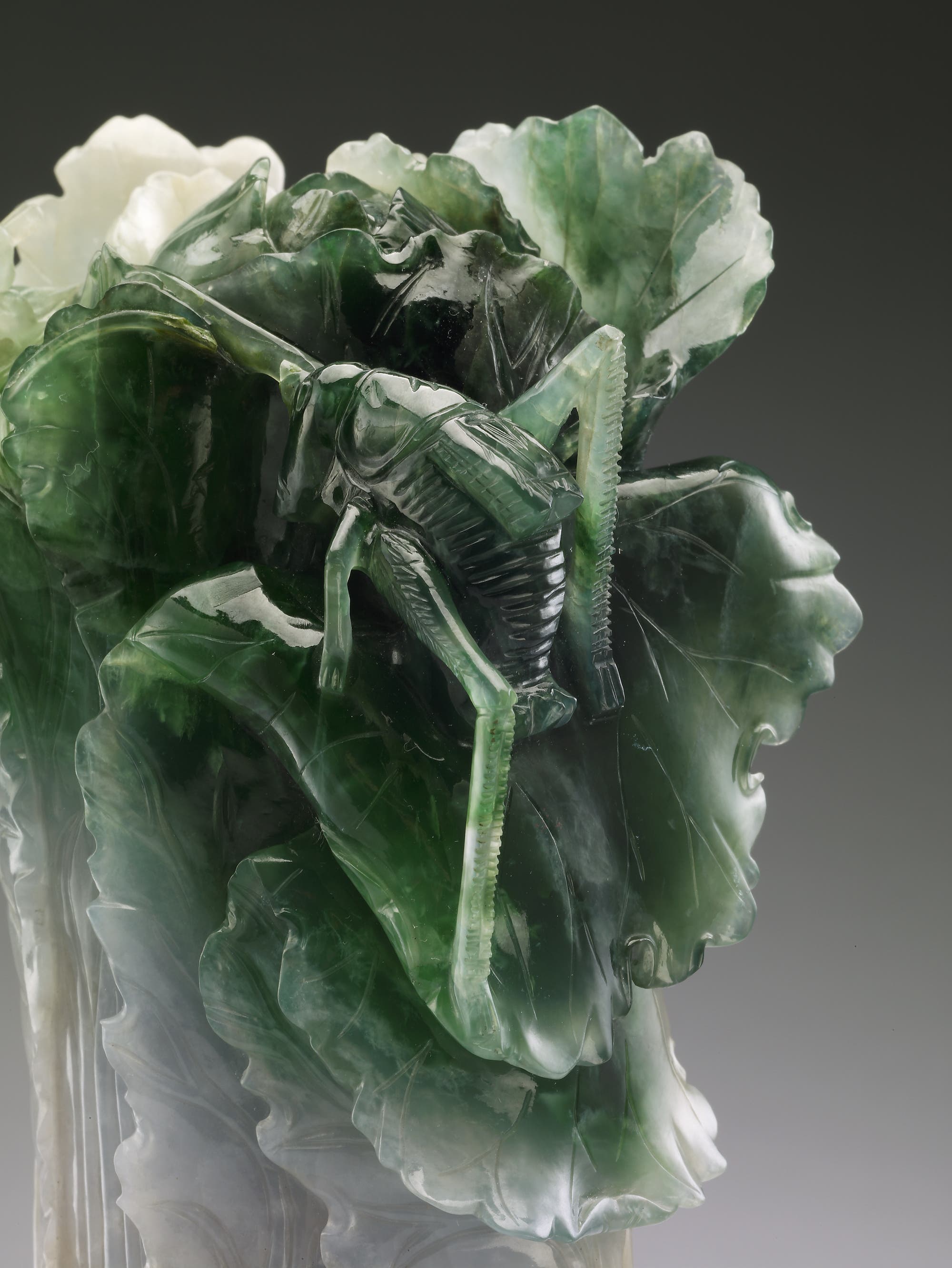 Eine kunstvolle Skulptur aus grünem und weißem Jadeit, die einen Kohlkopf mit einem darauf sitzenden Insekt darstellt. Die detaillierte Darstellung zeigt die feinen Blattstrukturen des Kohls und die präzise geschnitzten Merkmale des Insekts, was die handwerkliche Meisterschaft und die natürliche Ästhetik der Skulptur betont.