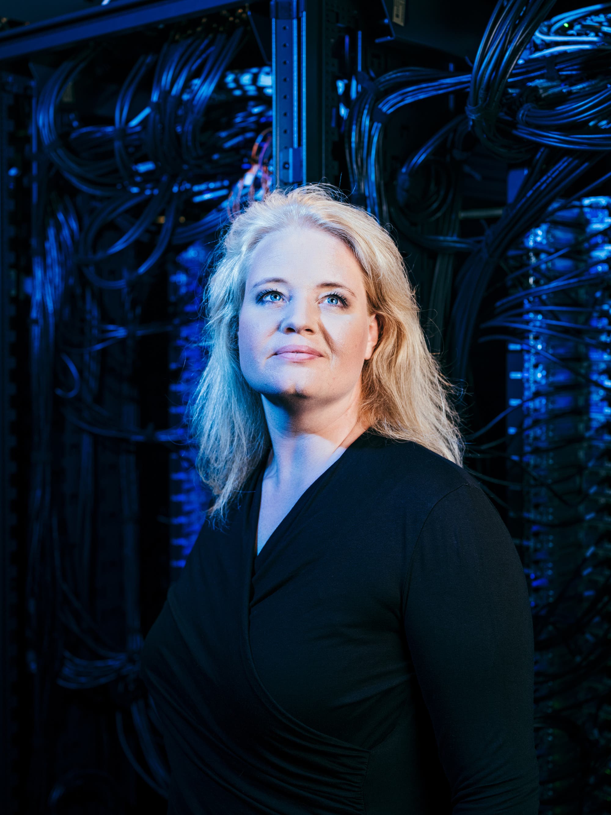 Eine Person steht in einem Serverraum, umgeben von zahlreichen Kabeln und Server-Racks. Die Beleuchtung ist in Blau- und Schwarztönen gehalten, was eine technische und futuristische Atmosphäre schafft. Die Person blickt nachdenklich in die Ferne.