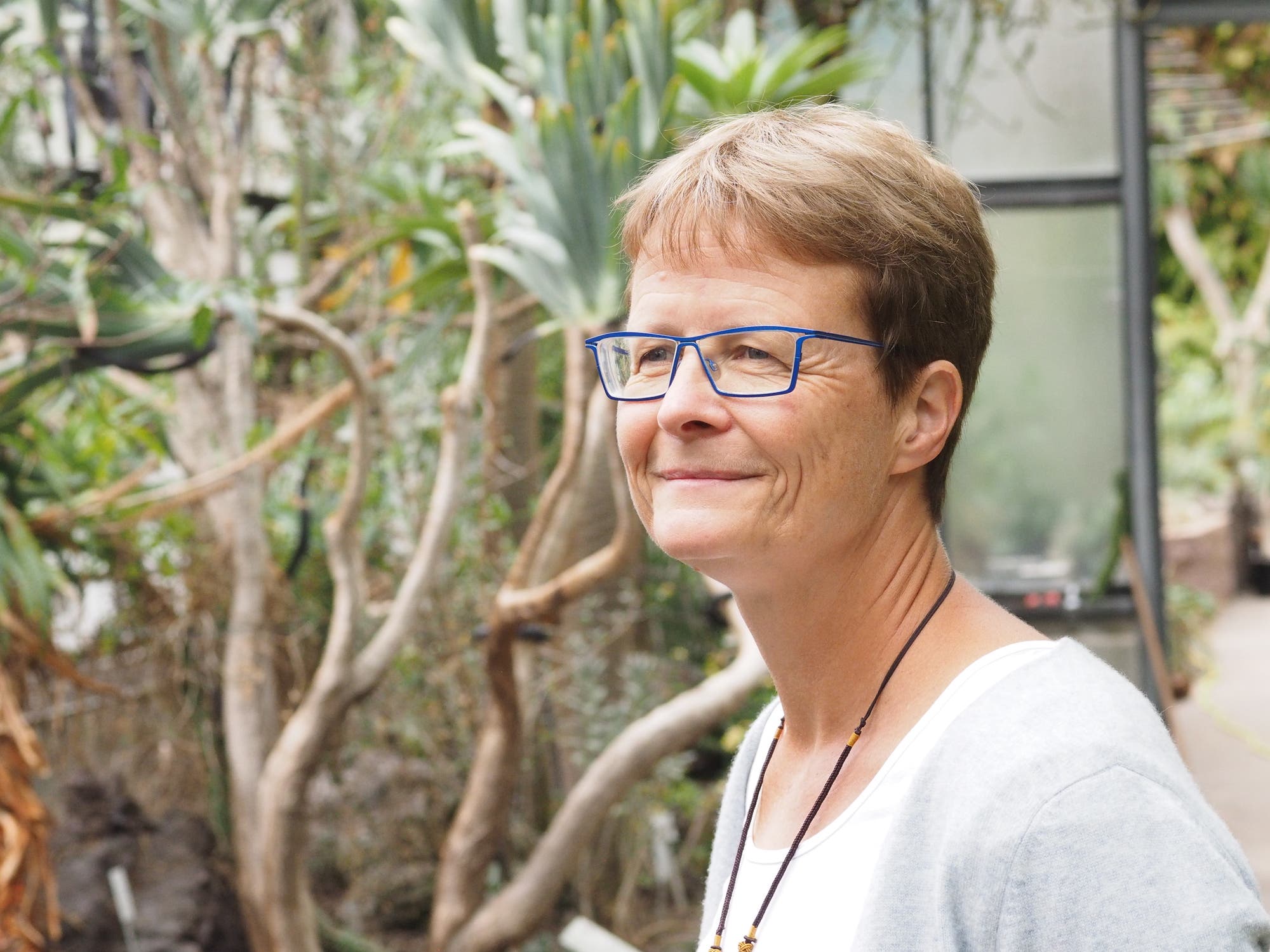 Eine Person mit blauer Brille steht in einem botanischen Garten. Sie trägt ein weißes Oberteil und eine graue Strickjacke. Im Hintergrund sind grüne Pflanzen und Bäume zu sehen. Die Person blickt lächelnd zur Seite.