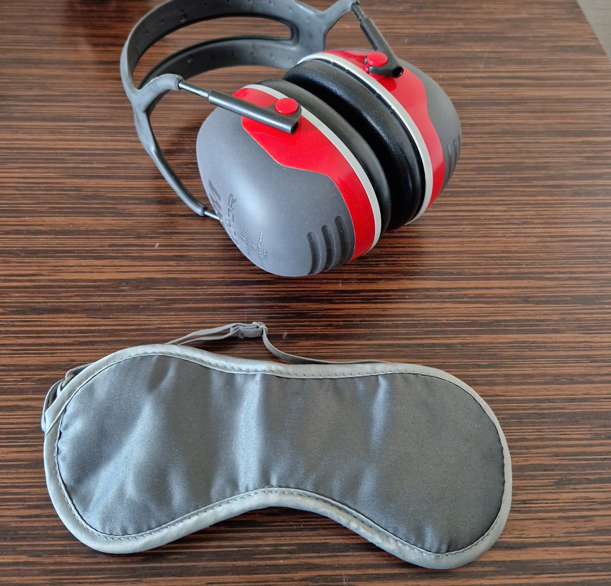 Ein Paar rot-schwarze Gehörschutzkopfhörer liegt auf einem Holztisch neben einer silbergrauen Schlafmaske. 