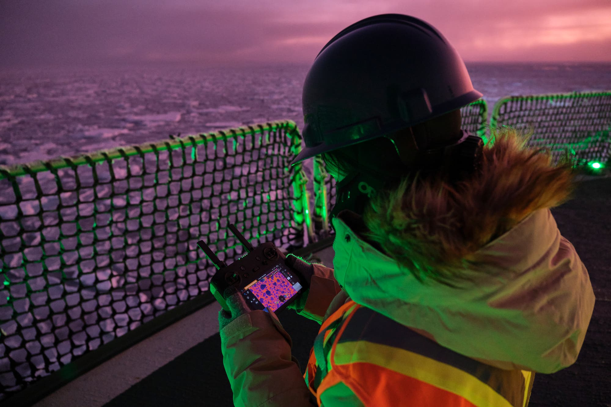 Eine Person in Schutzkleidung und Helm steht auf einem Schiff und bedient eine Fernsteuerung mit einem Bildschirm. Der Hintergrund zeigt eine eisige Landschaft bei Sonnenuntergang. Die Person trägt eine orange Sicherheitsweste und konzentriert sich auf die Steuerung. Die Szene vermittelt eine Atmosphäre von Kälte und Technologieeinsatz in einer extremen Umgebung.