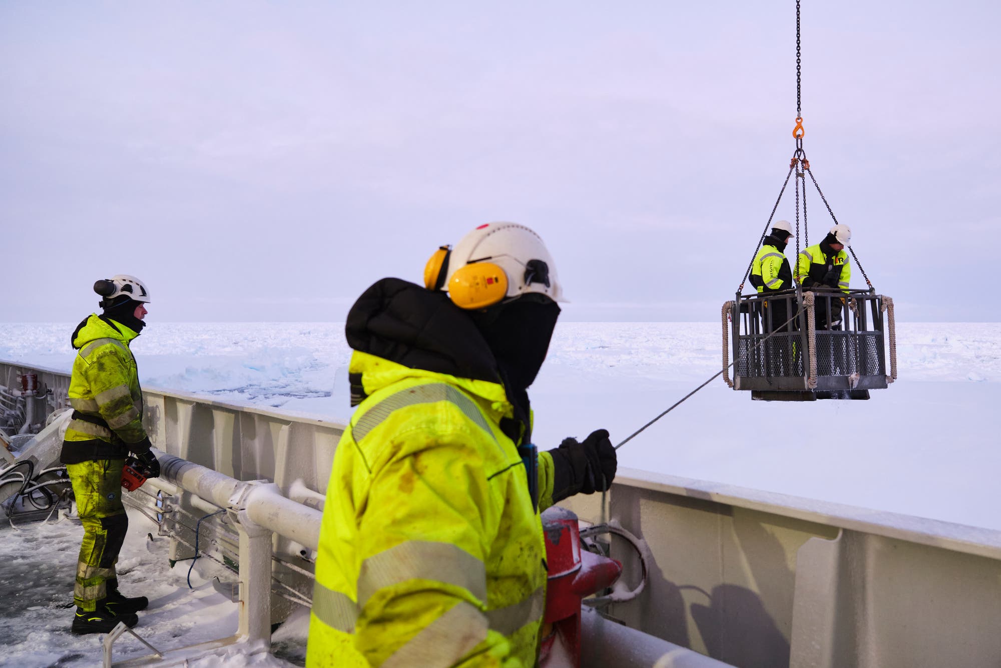 Personen in leuchtend gelber Schutzkleidung und Helmen arbeiten auf einem Schiff im Eis. Zwei Personen stehen in einem Metallkorb, der von einem Kran über das Eis gehoben wird. Eine weitere Person hält ein Seil, während eine vierte Person am Rand des Schiffs steht und in die Ferne blickt. Die Szene spielt in einer eisigen, arktischen Umgebung.