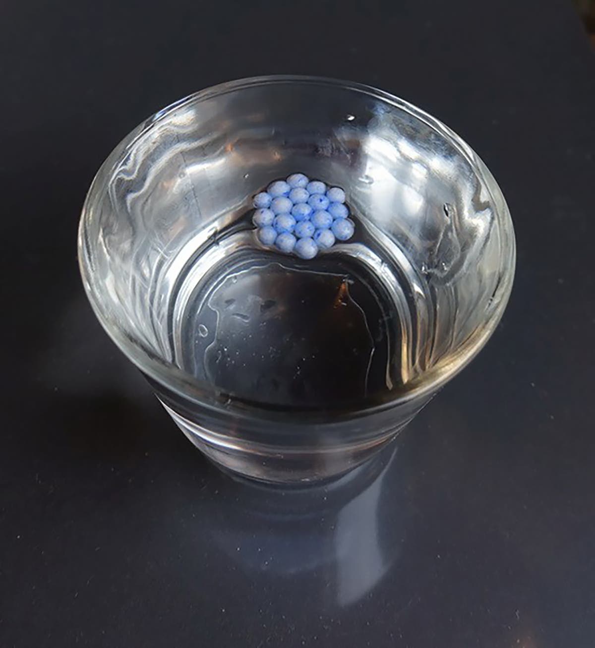Auf einer konvex über einem Trinkglas aufgewölbten Wasseroberfläche driften Styroporkügelchen zur Mitte