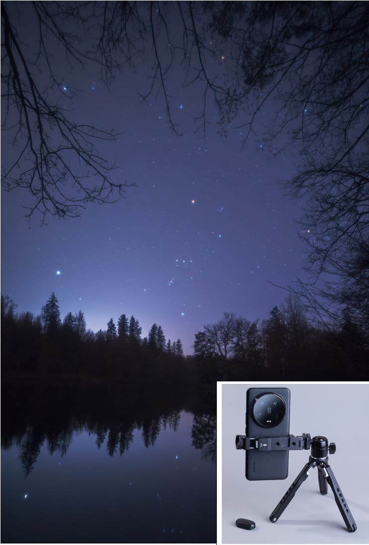 Eine nächtliche Landschaft mit einem klaren Sternenhimmel, der sich in einem ruhigen See spiegelt. Silhouetten von Bäumen umrahmen den Himmel. Im unteren rechten Bereich ist ein kleines Bild eines Smartphones auf einem Stativ zu sehen, das für die Astrofotografie verwendet wird. Der Himmel zeigt zahlreiche Sterne, die in der Dunkelheit leuchten.