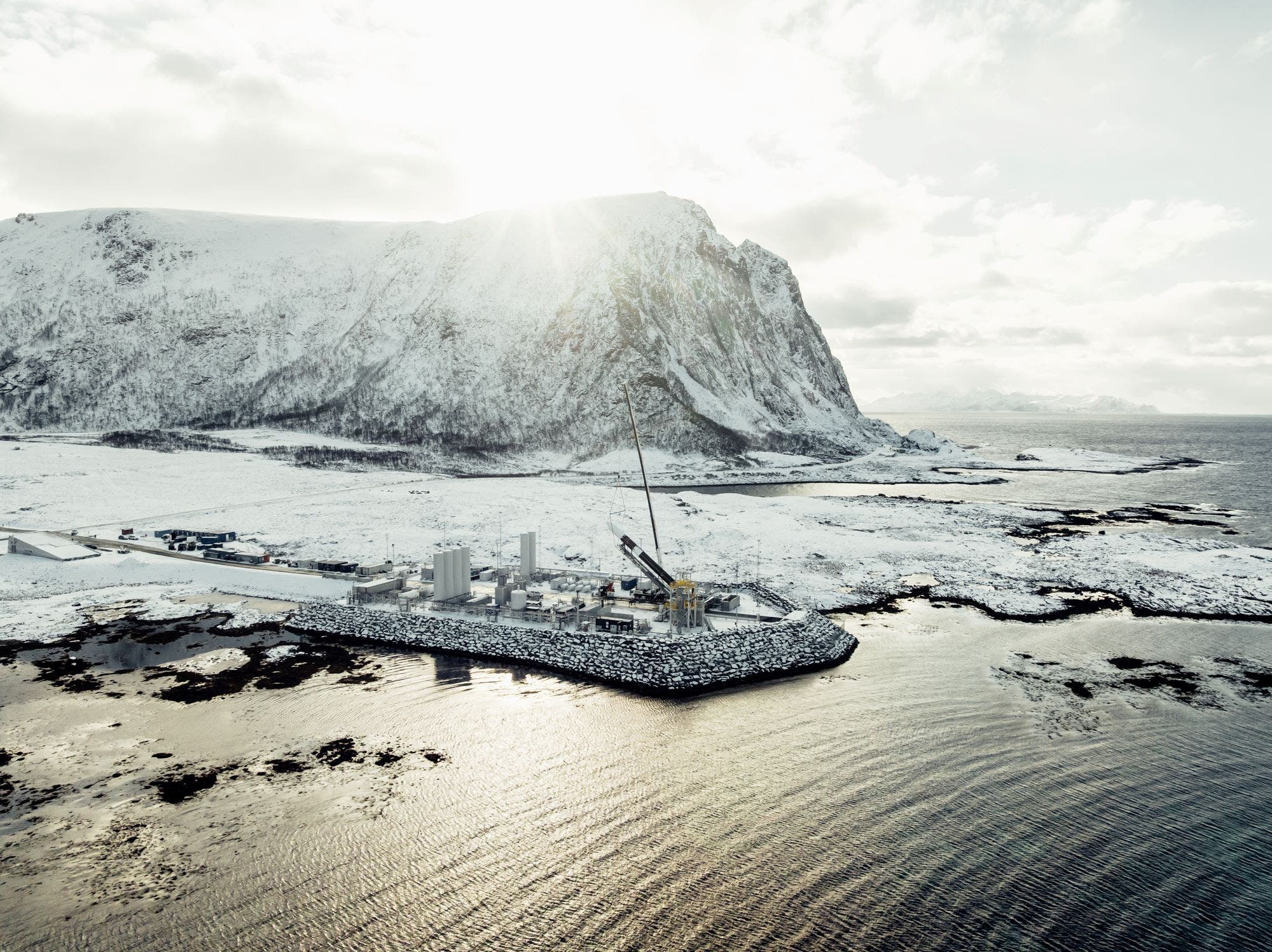 Eine verschneite Küstenlandschaft mit einem großen Berg im Hintergrund. Im Vordergrund befindet sich der Raketenstartplatz Andøya Space Center auf einer Landzunge, die ins Meer ragt. Ein Kran ist sichtbar, der auf dem Gelände arbeitet. Die Sonne scheint hinter dem Berg hervor und beleuchtet die Szene. Das Bild vermittelt eine abgelegene, winterliche Umgebung.